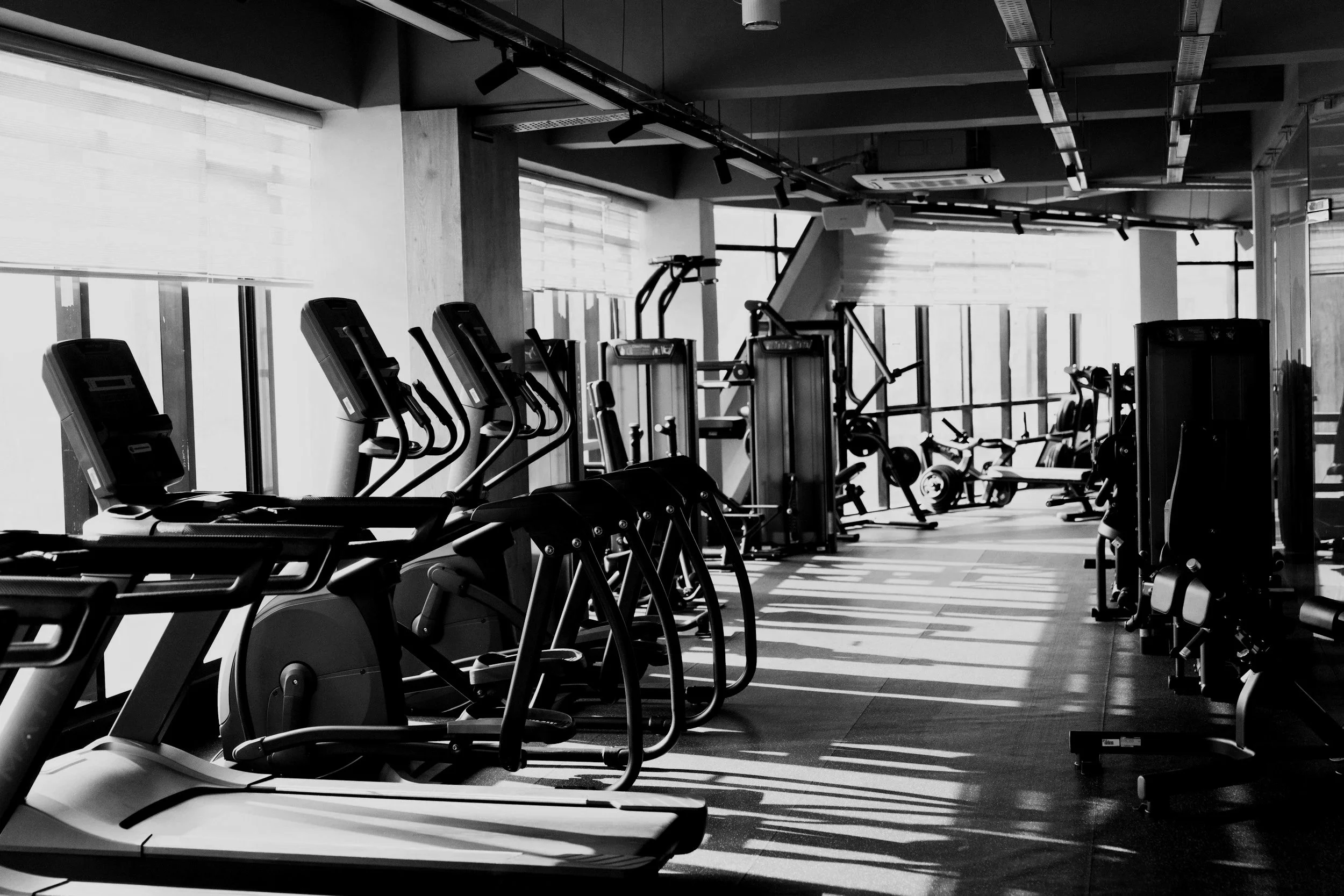 A black and white photo of a modern gym with treadmills and other workout equipment near large windows.