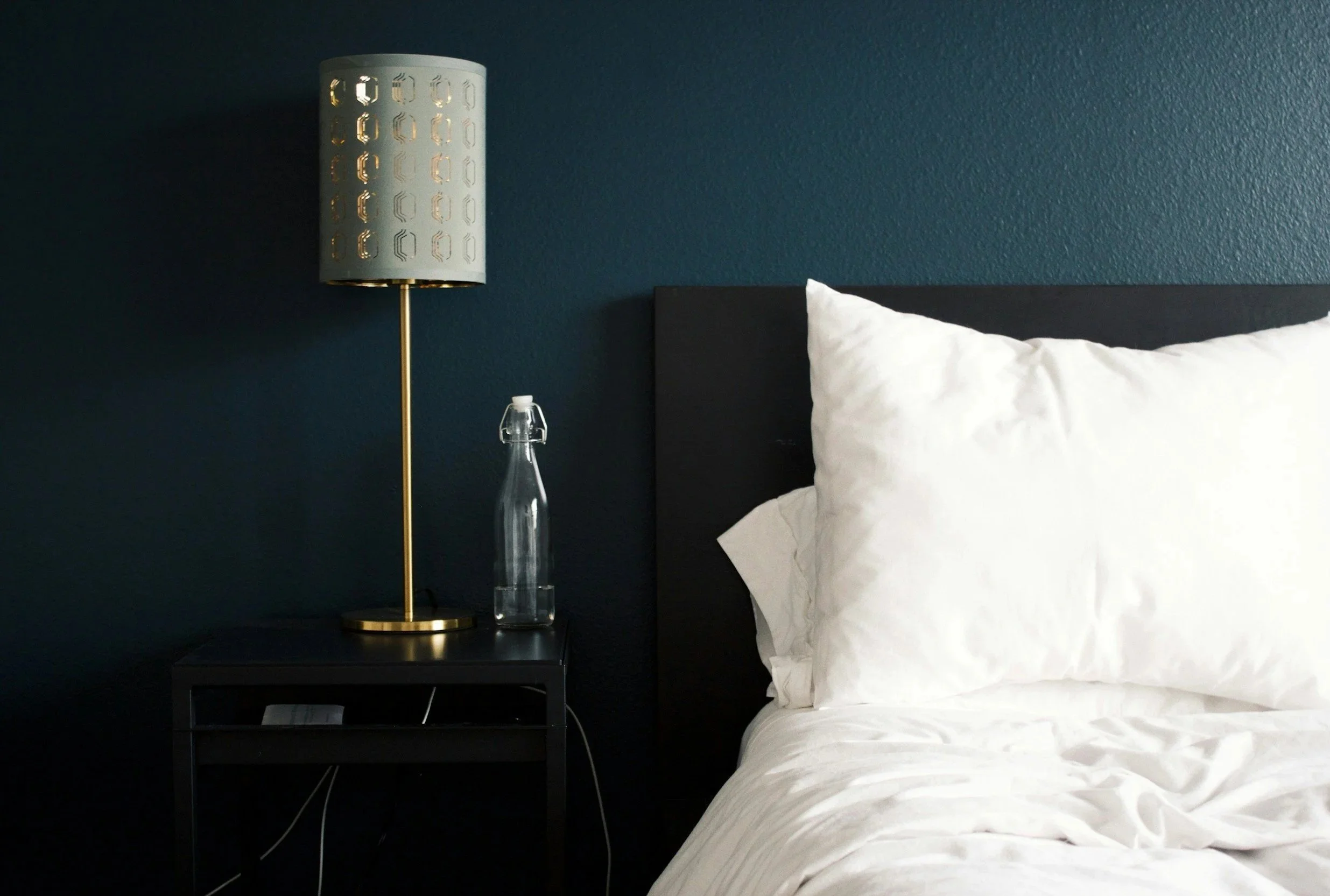A bedroom with a navy blue wall, a black bed frame with white bedding, a black nightstand, a modern white and gold table lamp, and a glass water bottle on the nightstand.