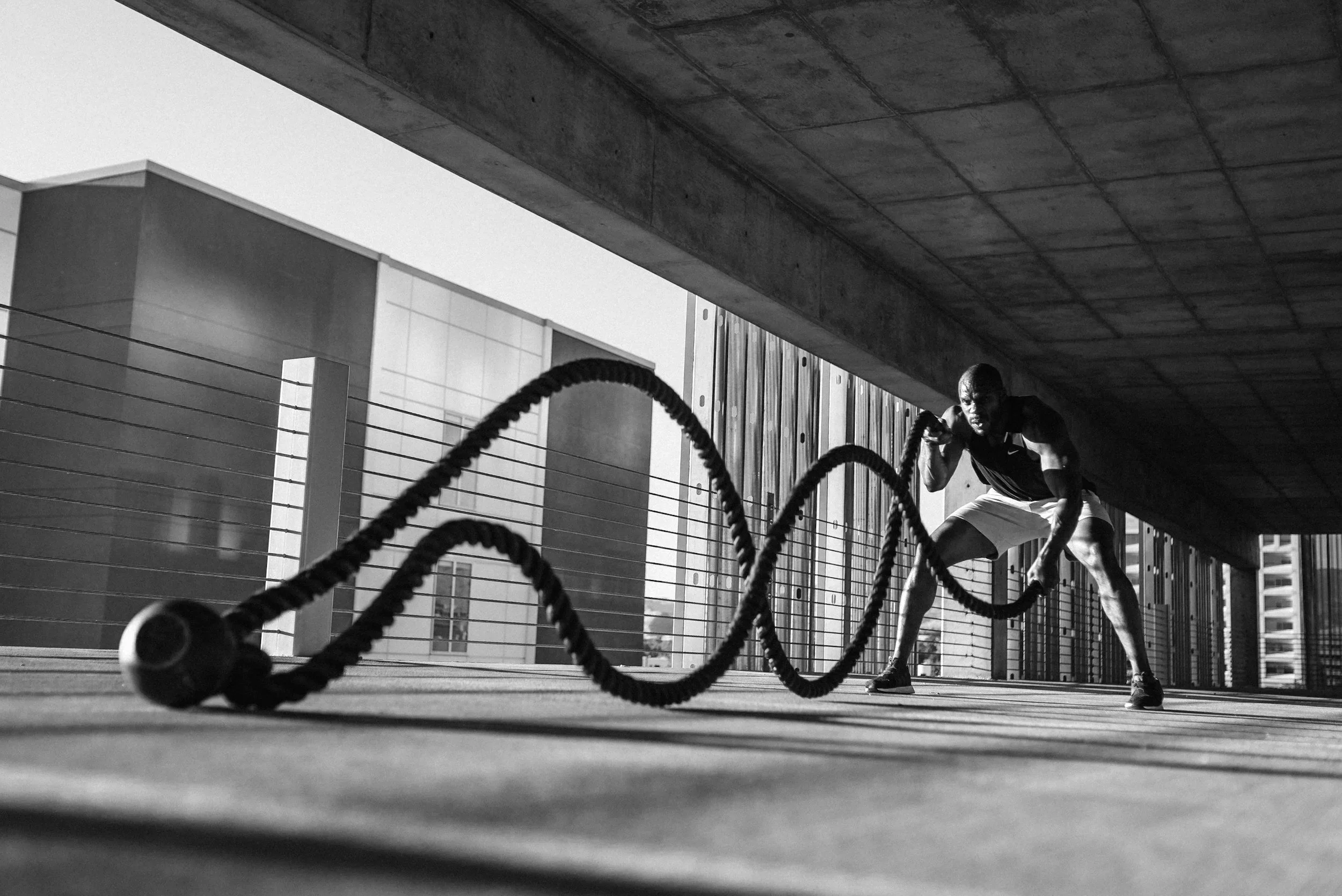 A man in athletic attire is exercising outdoors under a bridge, pulling on thick battle ropes for strength training.