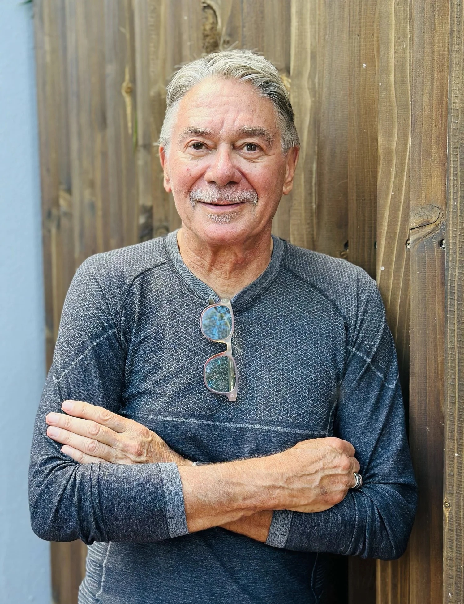 Older architect in a gray shirt with folded arms standing against a wooden wall, wearing glasses around his neck.