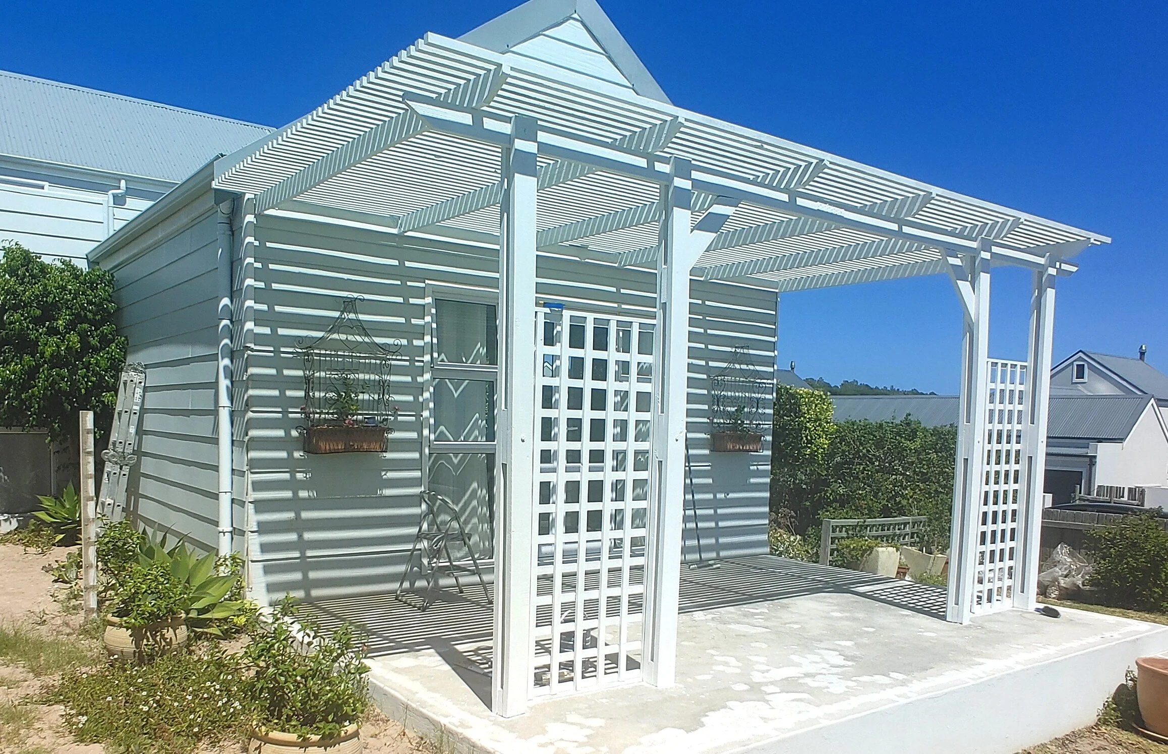 White pergola attached to a small building with light blue siding, surrounded by a garden with green plants and a clear blue sky.