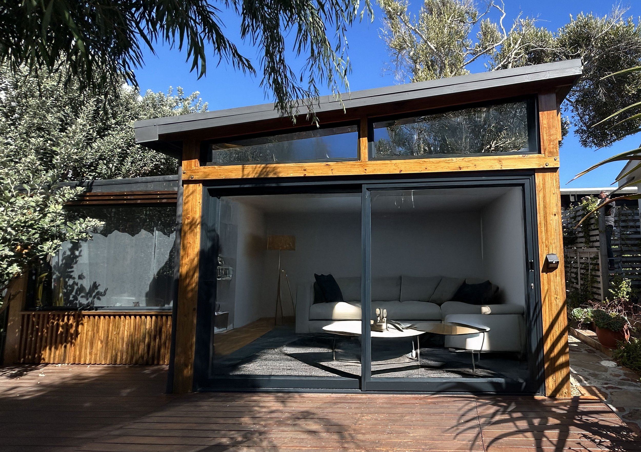 A modern small house with a wooden exterior and large glass sliding doors, revealing a cozy interior with a couch and coffee table. The outdoor area features wooden decking and surrounding greenery under a clear blue sky.