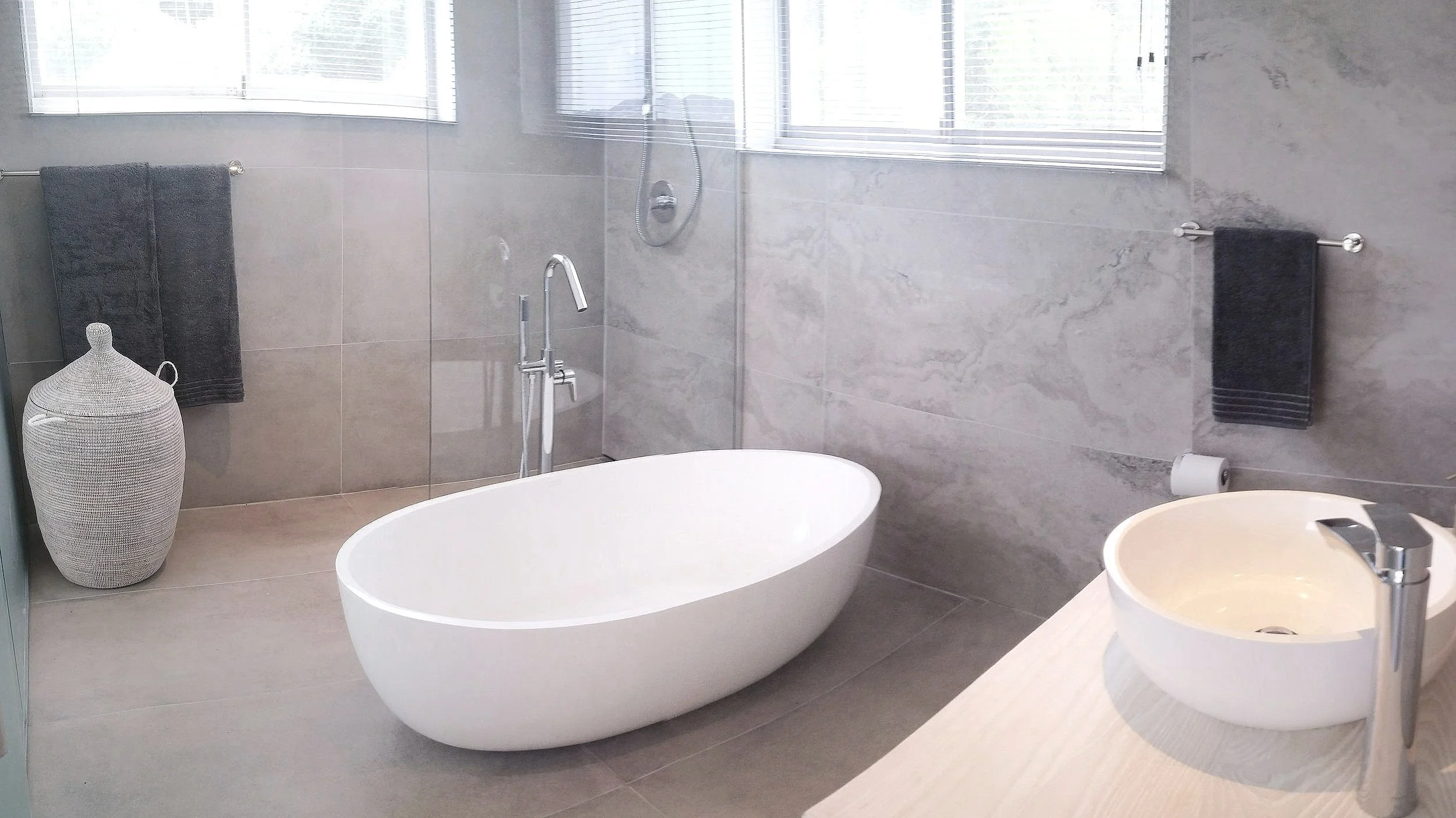 Modern bathroom with a freestanding white bathtub, a round ceramic sink on a countertop, chrome fixtures, a woven basket, and gray towels on towel racks. The room has gray marble walls and a large window with blinds.