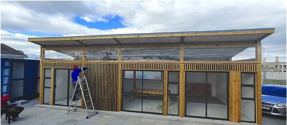 A construction worker on a ladder next to a modern wooden building with large glass windows and a corrugated roof.