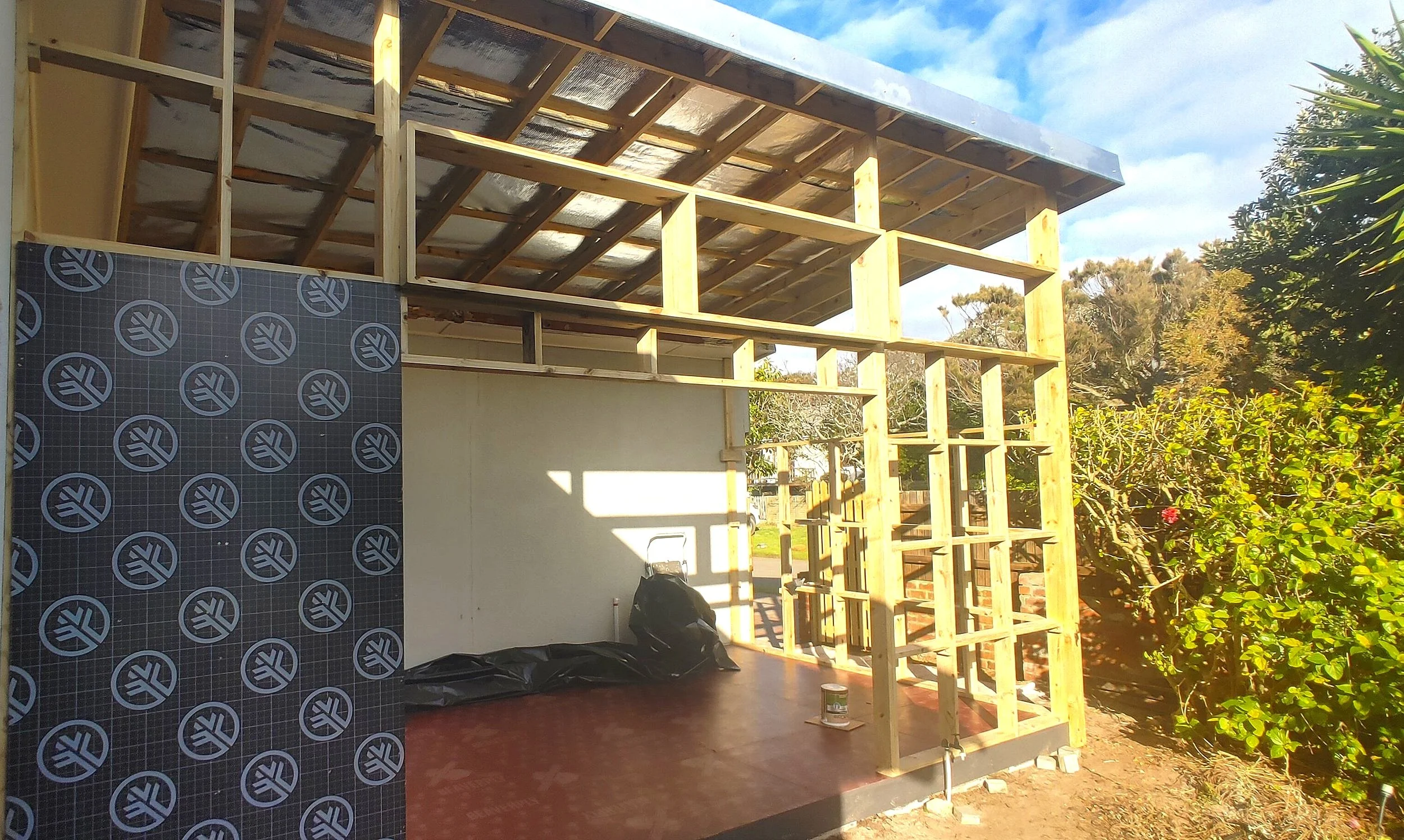 Partially constructed wooden frame of a small building with some insulation and roofing in place, surrounded by greenery.