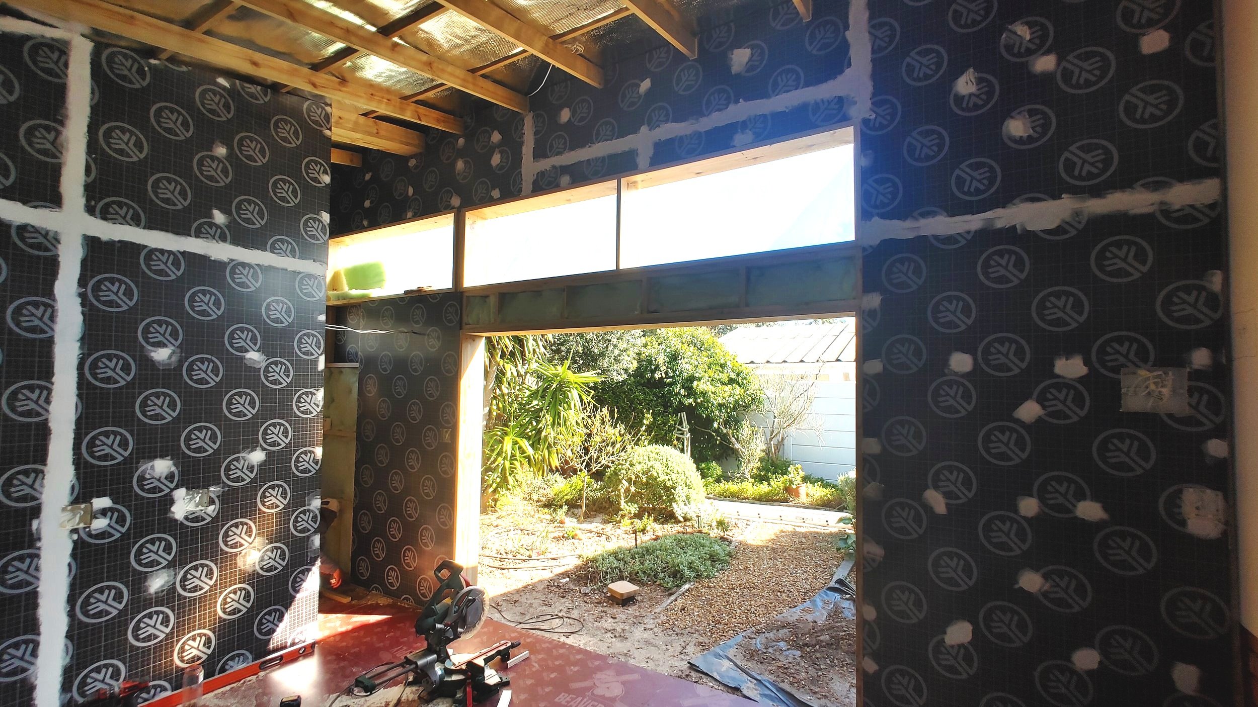Interior of a building under construction with dark insulation panels on the walls and an opening leading to a garden outside.