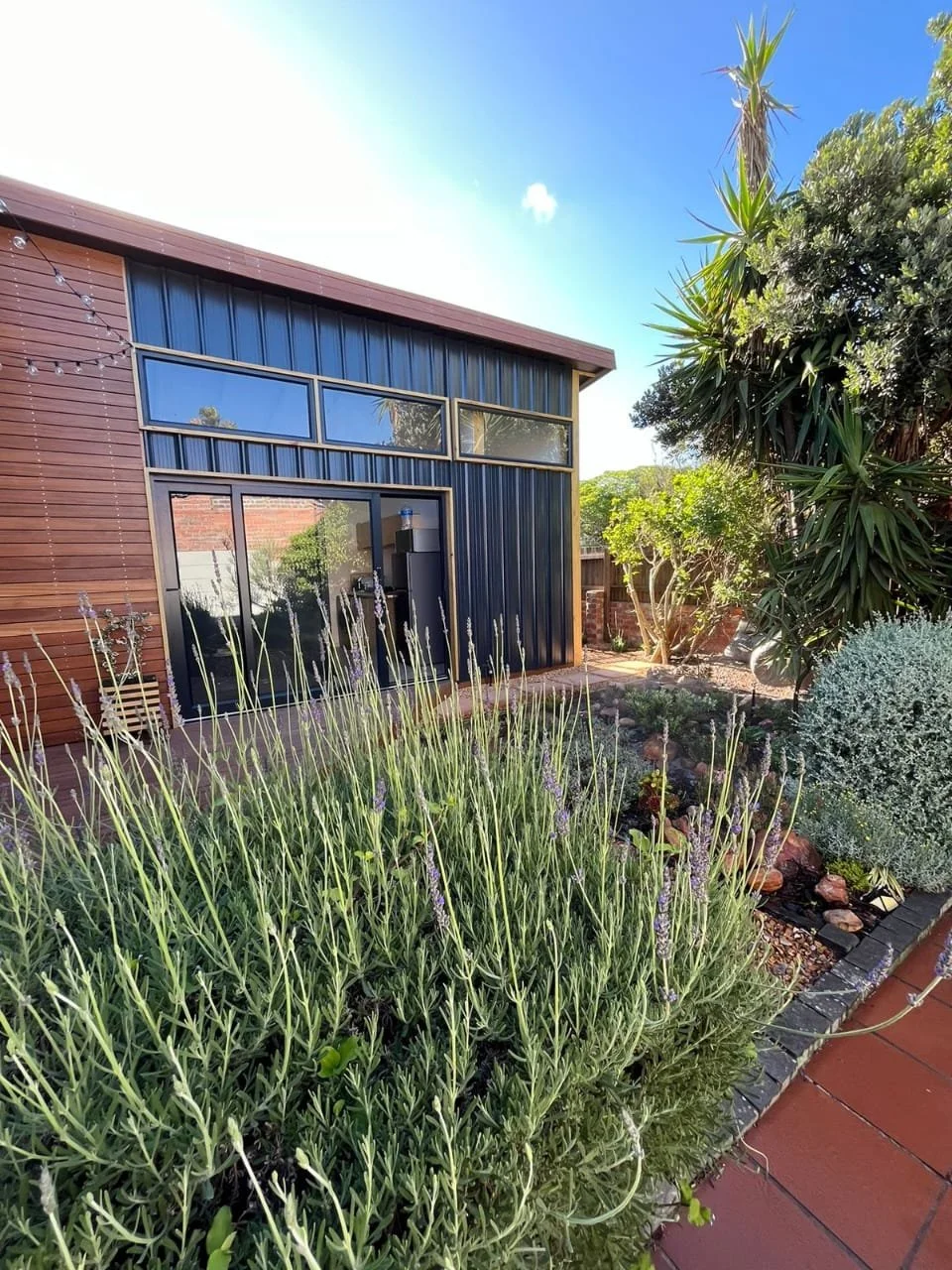 Modern house with wood and metal exterior, large windows, garden plants, and a clear blue sky.