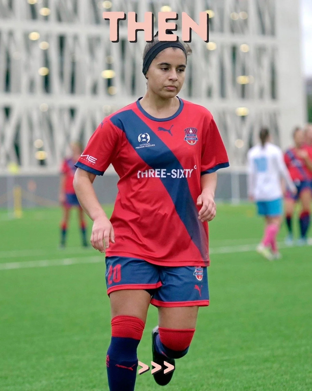 A female soccer player in a red and blue uniform walking on a soccer field during a game. She is wearing a black headband and has a focused expression. The background shows other players and a modern building.
