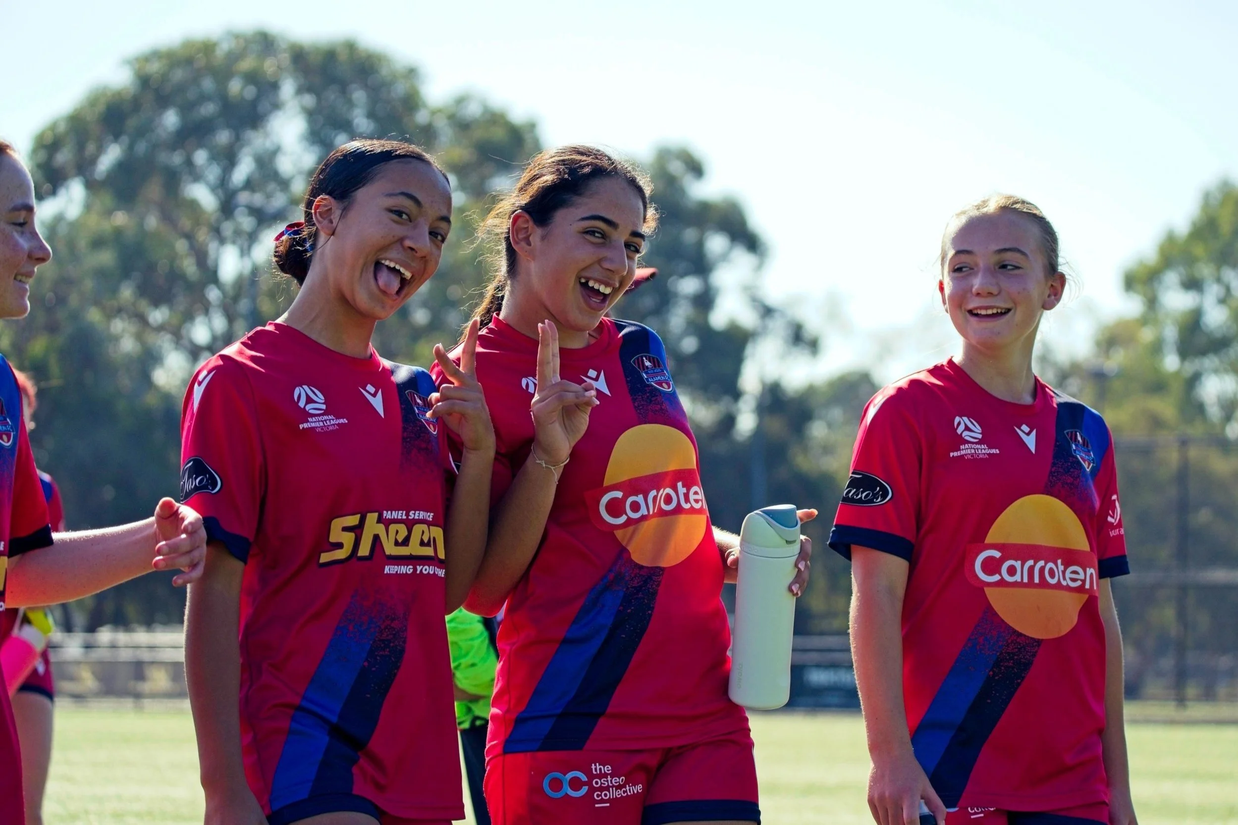 Four young female soccer players wearing red jerseys with sponsorship logos standing outdoors on a sunny day, smiling and gesturing.