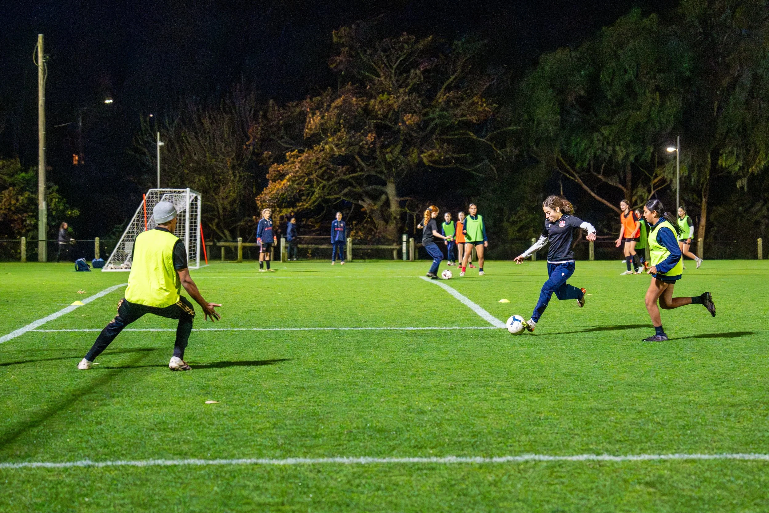 A group of people playing soccer on a well-lit field at night. Some players are in green vests, others in black and orange, with a few in regular sportswear. One person is about to kick the ball, while a goalkeeper is in a ready position near the goal, and others are watching or preparing for play. Trees are visible in the background.