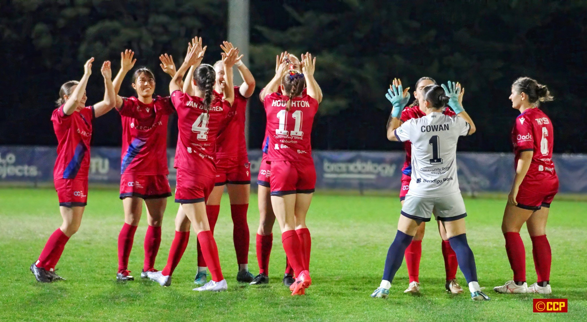 Women soccer team celebrating on the field during a match at night. The players are wearing red uniforms, with one in a white goalkeeper jersey facing them. They are clapping and high-fiving each other.