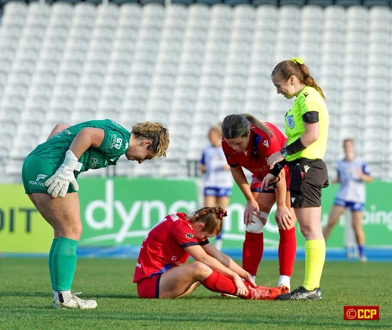 A female soccer player is sitting on the grass, holding her ankle, appearing injured after a game. Two other women, one possibly a teammate and another in a referee uniform, are attending to her on the field. A goalkeeper in a green uniform looks on. In the background, other players and empty stadium seats are visible.