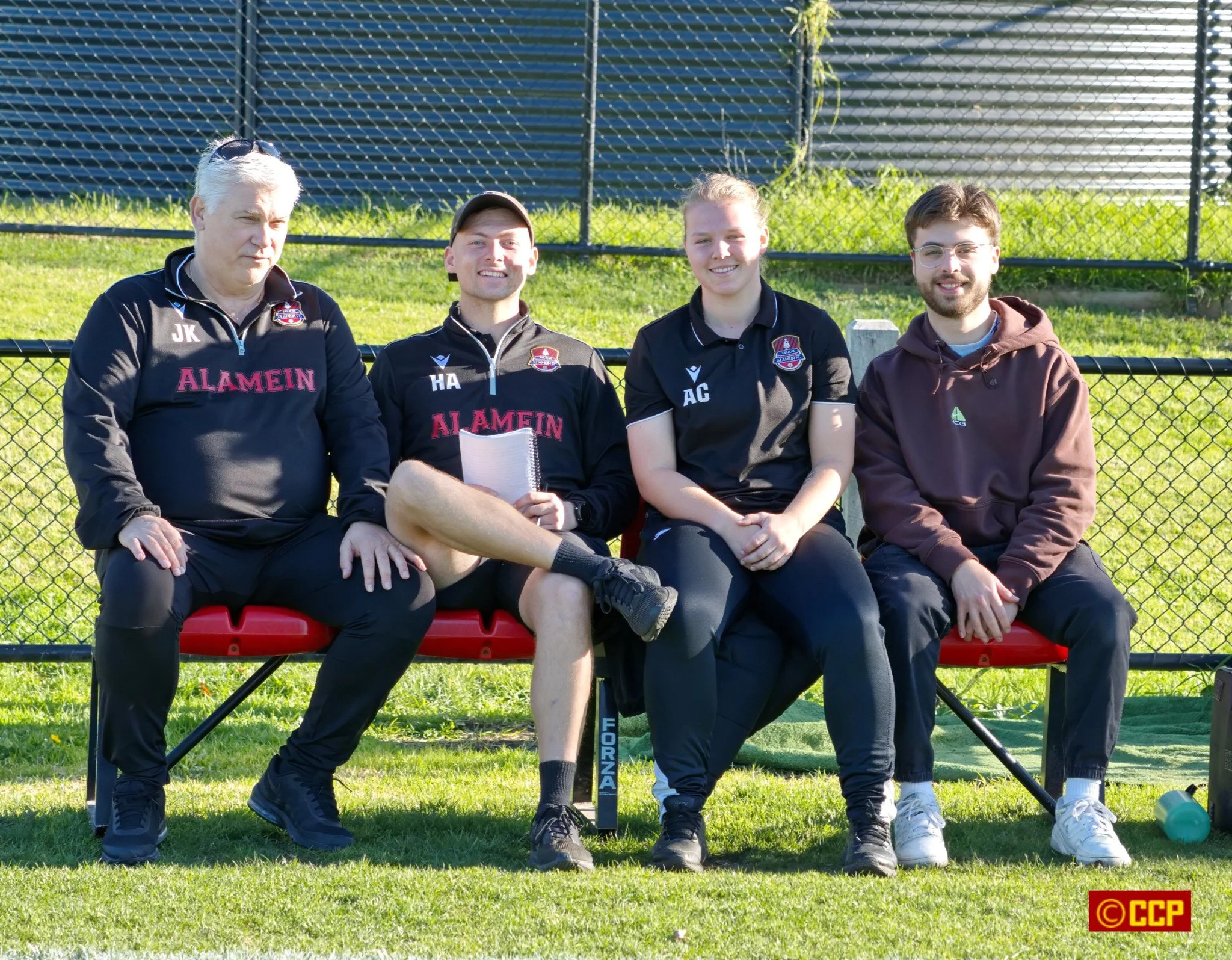 Four people sitting on a bench outdoors, three wearing black sports jackets with 'ALAMEIN' written on them and one in a hoodie, all smiling, with a fence and grassy area in the background.