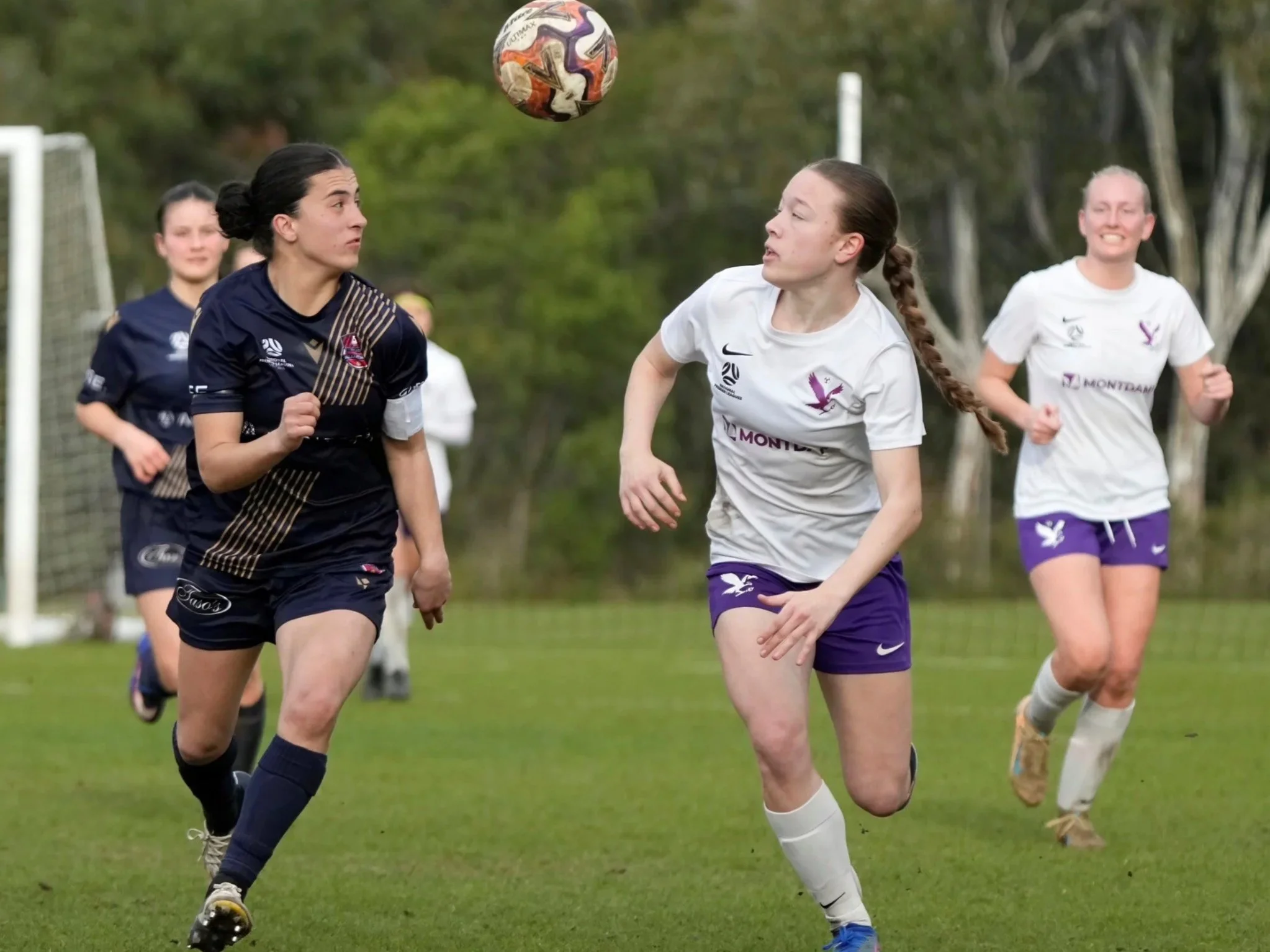 Women playing soccer on a field, one in a dark jersey and the other in a white jersey, chasing a colorful soccer ball.