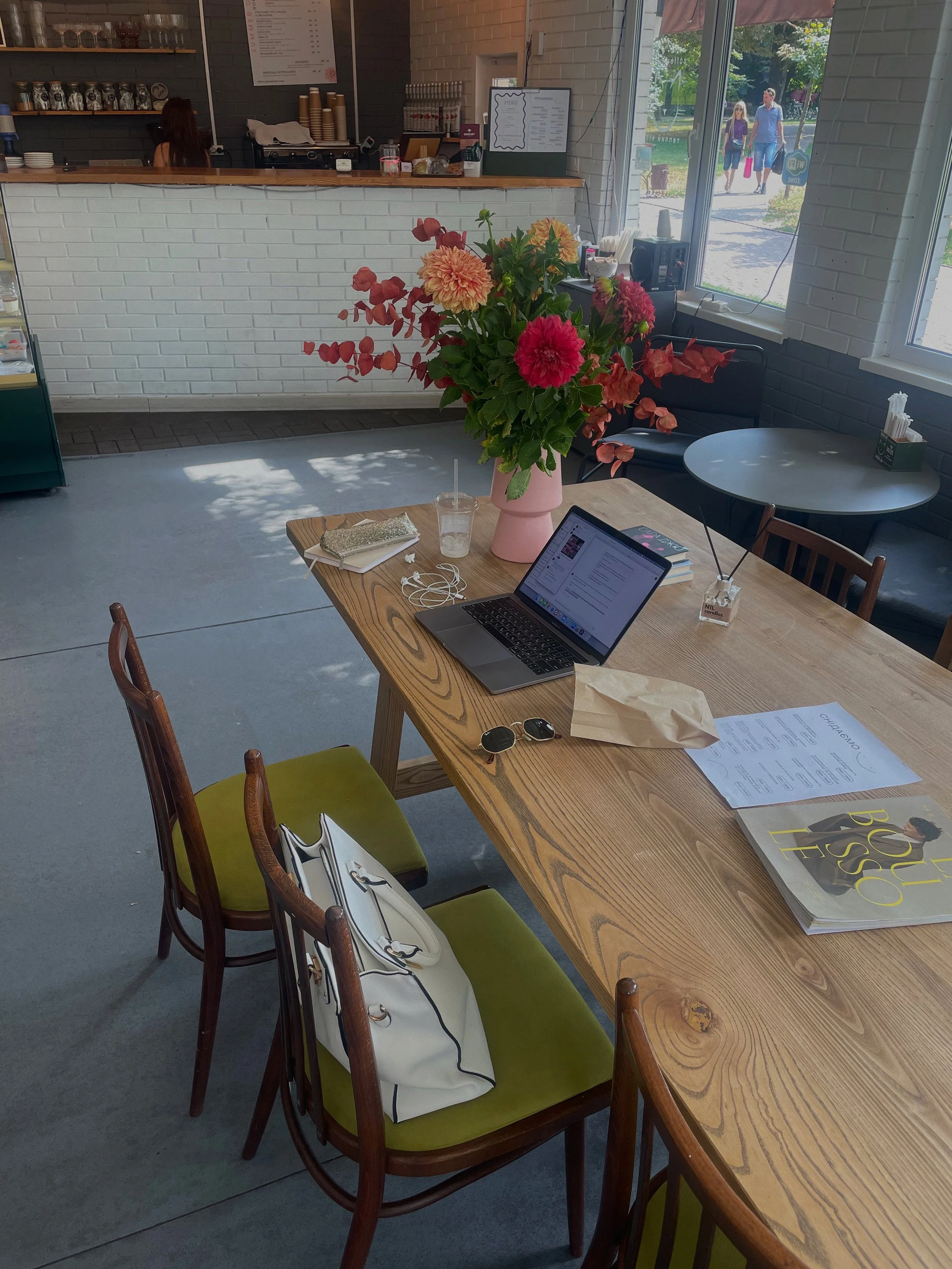A cozy cafe interior with a large wooden table, filled with a laptop, sunglasses, papers, and a white handbag. A vibrant flower arrangement in a pink vase is in the center of the table. In the background, a counter with a person, a menu on the wall, and large windows showing people outside, with trees and a sunny day.