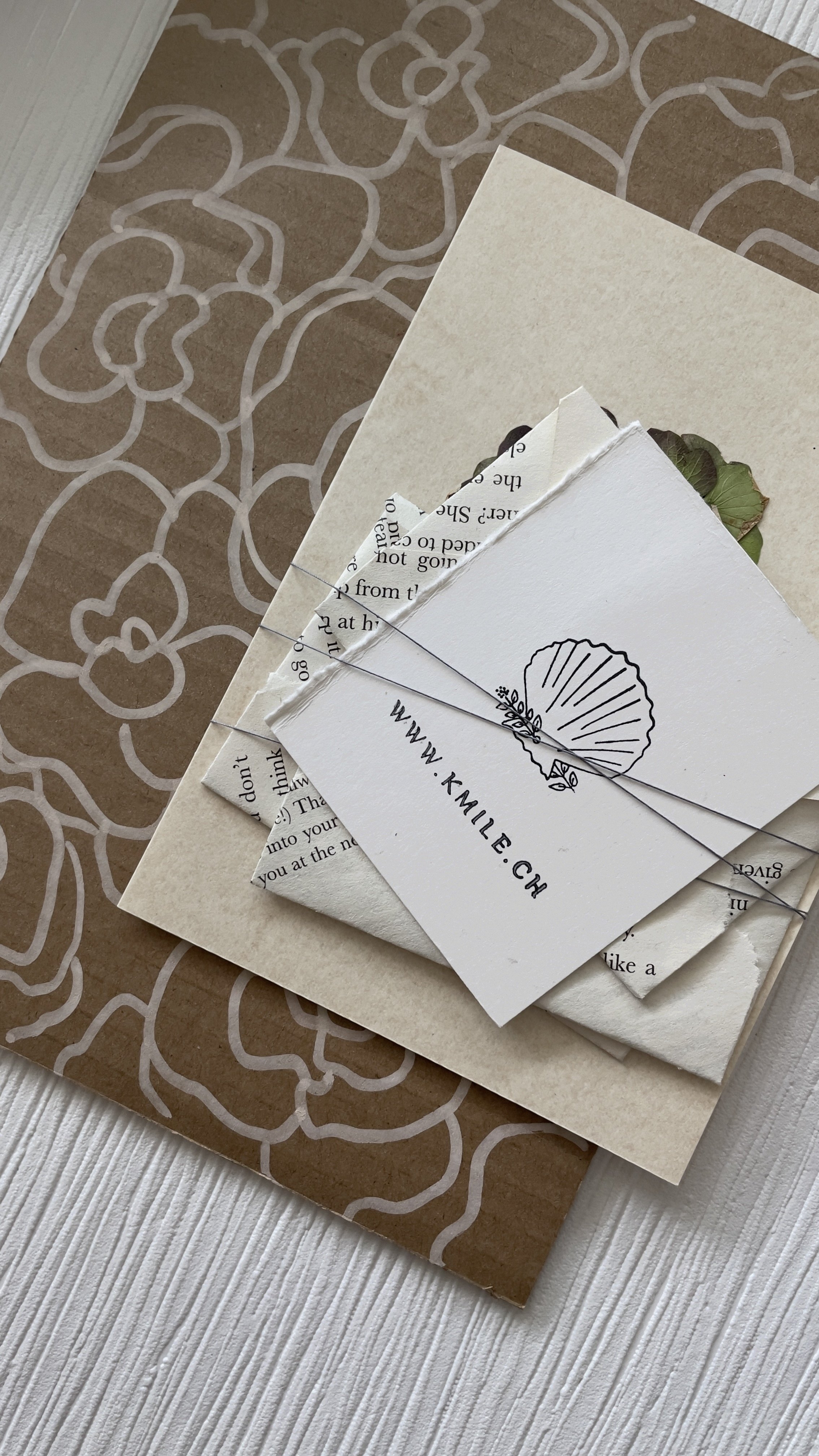 A collection of paper items, including a card with a shell illustration, a piece of torn newspaper, and a beige envelope, all arranged on a brown patterned surface with a floral design for branding