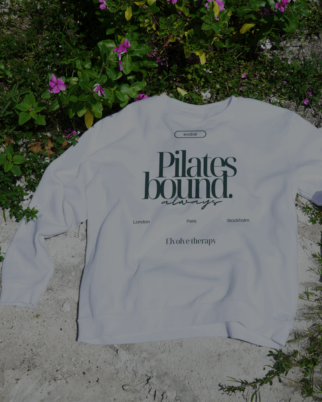 White sweatshirt with black text that reads 'Pilates bound. always' and mentions locations London, Paris, Stockholm, and brands Evolve therapy and evolve. The sweatshirt is laid on sandy ground with green plants and purple flowers around.