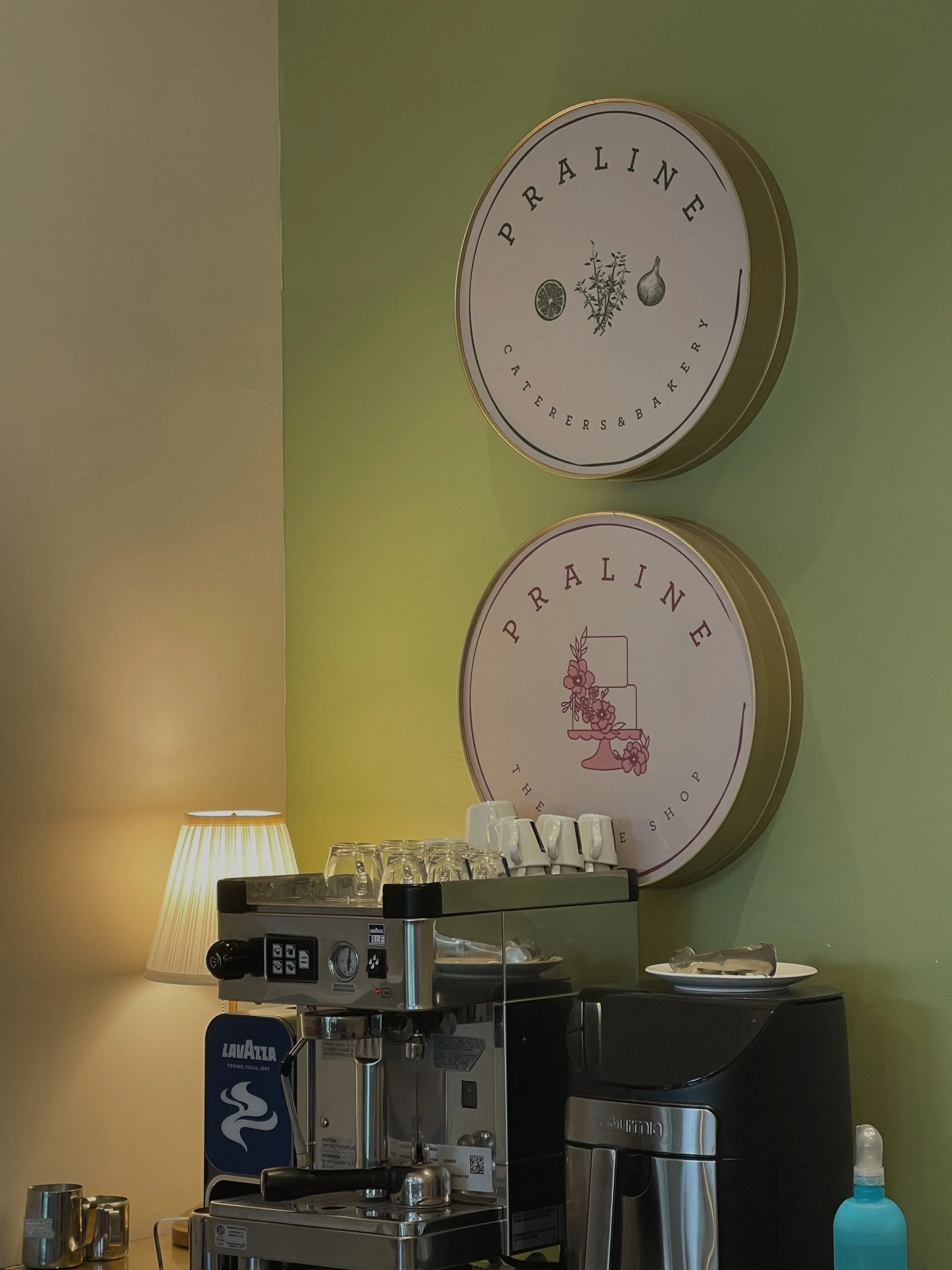 Coffee station in a cafe, with a La Marzocco espresso machine, cups, a thermal carafe, a small plate, a lamp, and two large round signs on a green wall reading "PRAISE Caterers & Bakery" and "PRAISE The One Stop Shop".