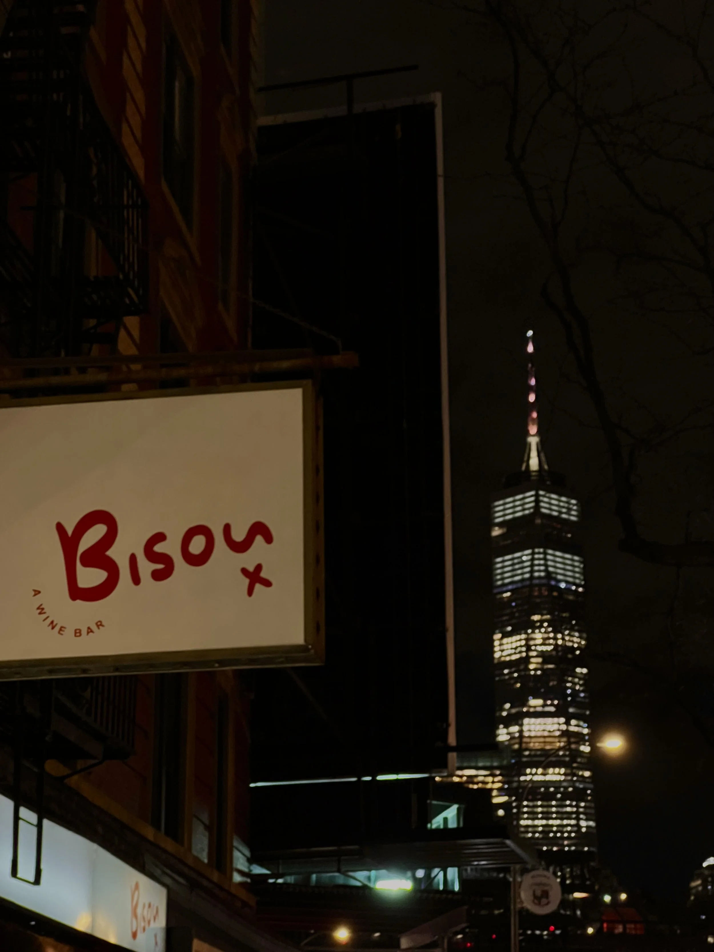 Nighttime cityscape with illuminated One World Trade Center in the background and a sign for a wine bar called Bisou in the foreground.