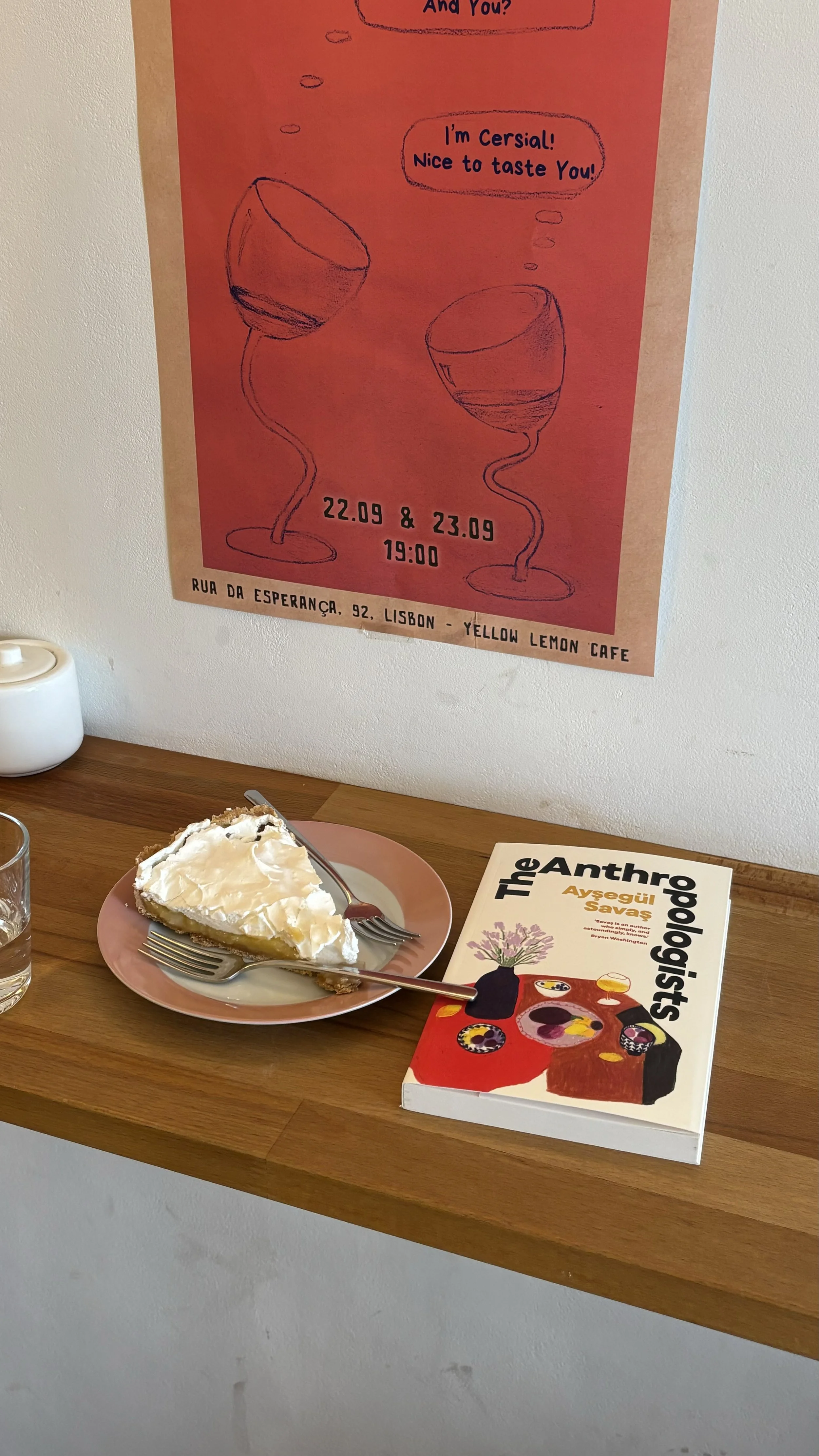 A slice of lemon meringue pie on a pink plate with a fork, a book titled 'The Anthropologists' by Ayşegül Savaş on a wooden table, a glass of water, a white teapot, and a poster on the wall behind with drawings of wine glasses and event details.