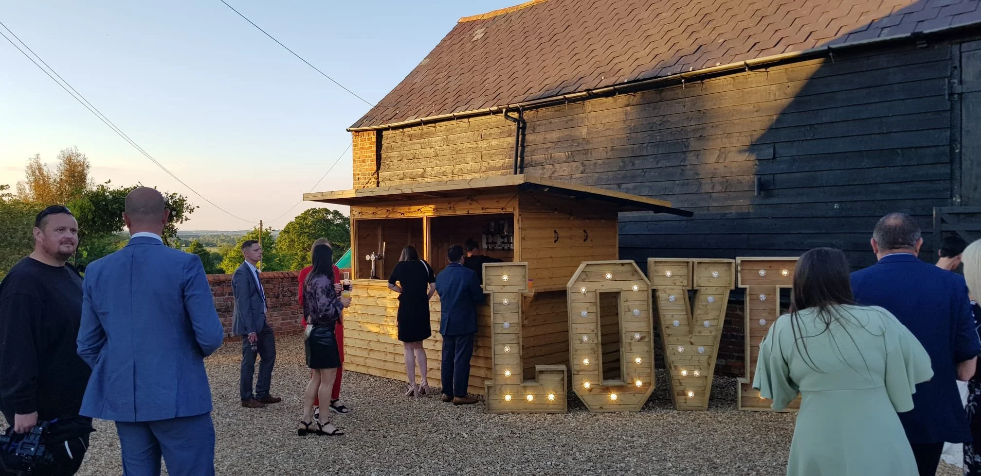 Outdoor event with people near a wooden bar and large illuminated letters spelling 'LOVE'