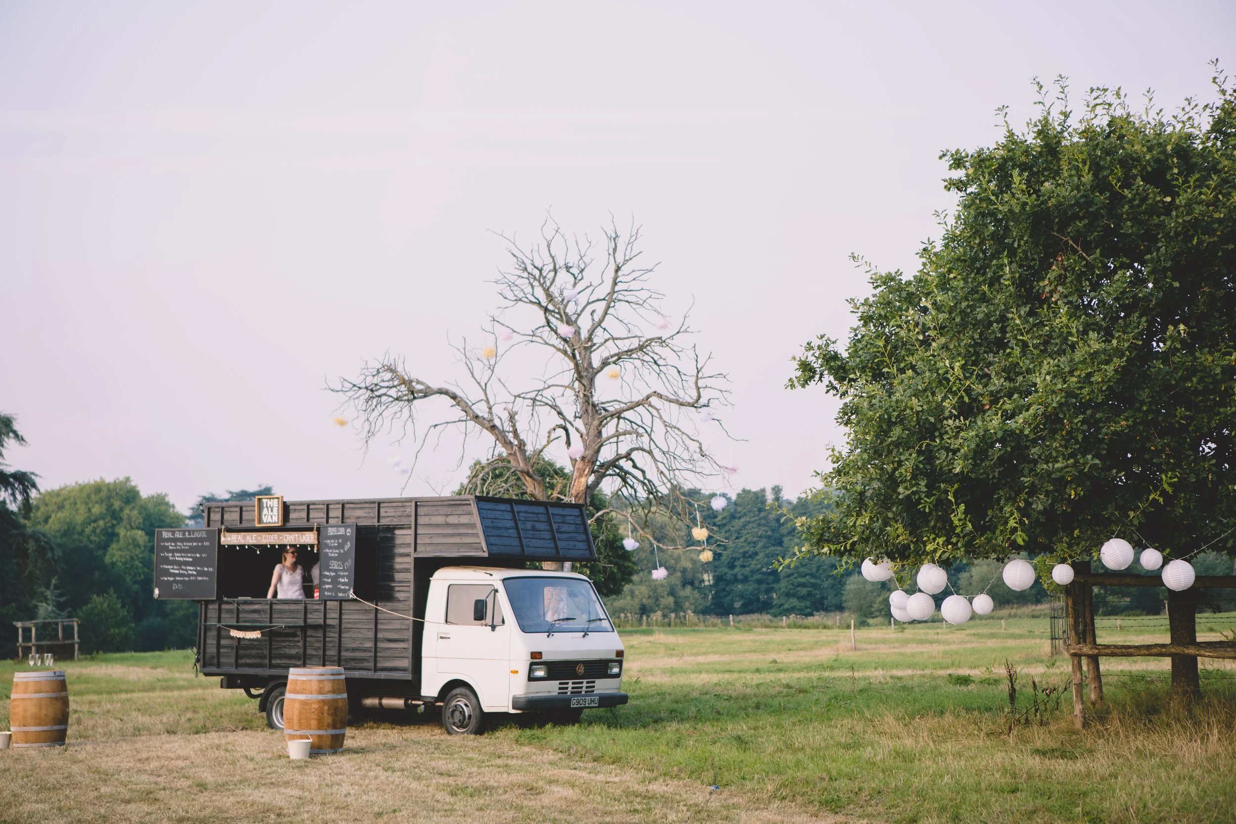A white food truck set up in a field with a black serving cabin, complemented by barrels and hanging lanterns on nearby trees.