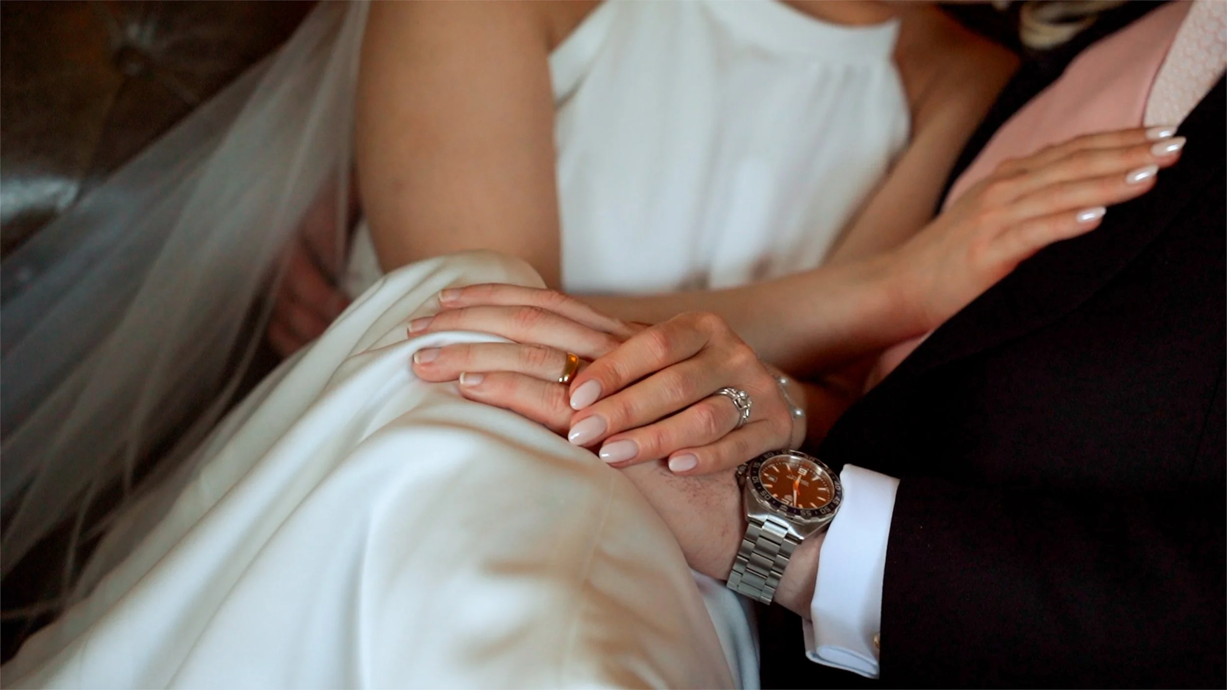 Close-up of a hand with wedding rings resting on a person's arm, with the person wearing a white shirt and another person dressed in dark clothing nearby.