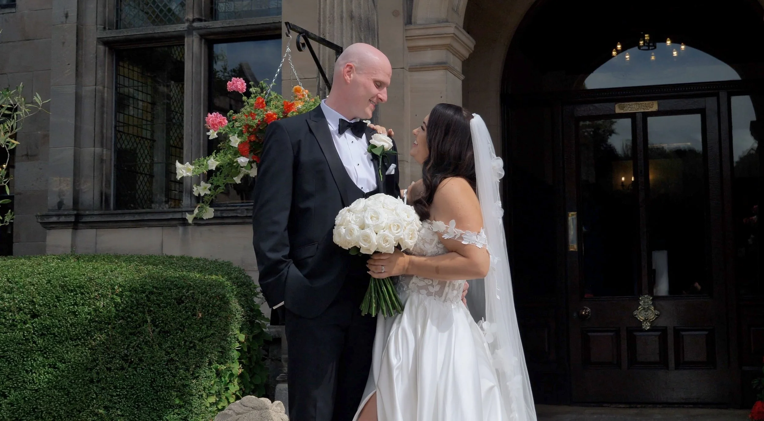 A bride and groom stand close together outside a building, smiling and gazing into each other's eyes. The bride is holding a bouquet of white roses, and the groom is wearing a black tuxedo with a bow tie. There are flowers behind them and an arched doorway in the background.