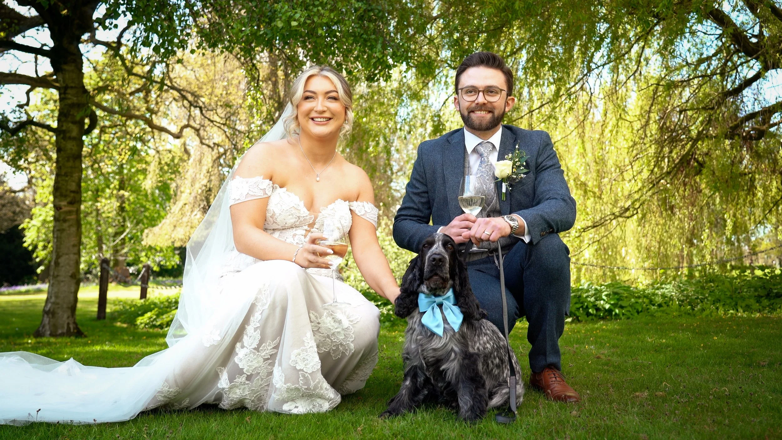 A bride and groom sitting outdoors in a park with green trees, smiling and holding drinks, accompanied by a black and white dog with a blue bow.