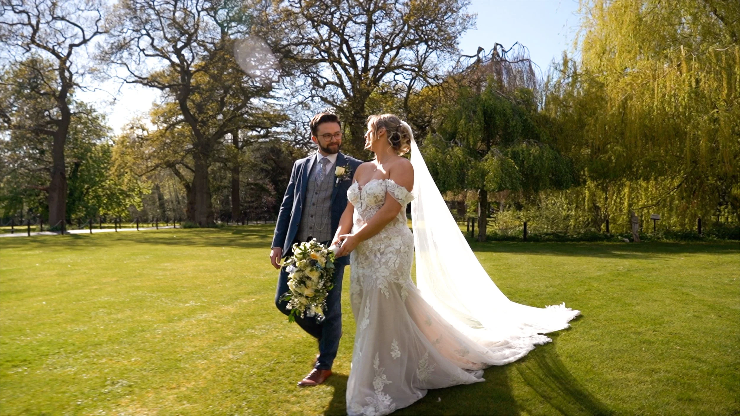 A bride and groom standing outdoors on a grassy area, holding hands and gazing at each other. The bride wears a white lace wedding gown with off-the-shoulder sleeves and a long veil, while the groom wears a blue suit with a vest and glasses. The brid