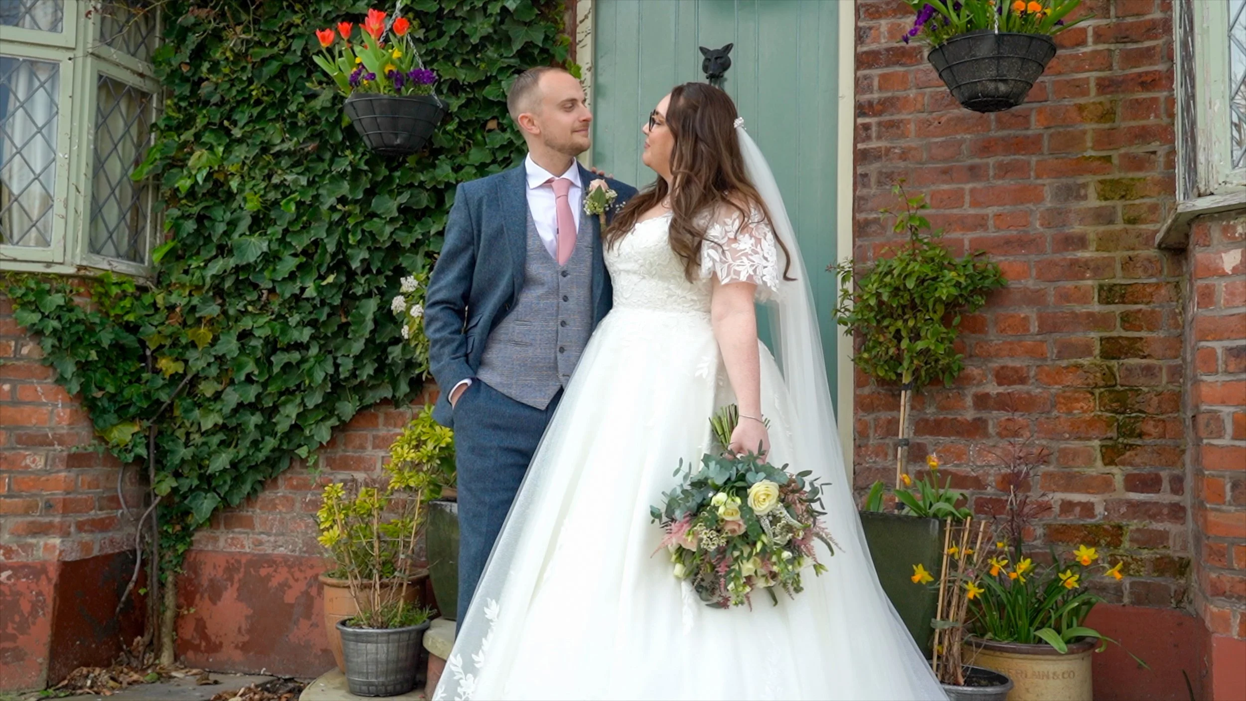 A bride and groom stand close together outside in front of a brick wall, looking at each other. The bride is wearing a white wedding dress with lace details and holding a bouquet of flowers. The groom is dressed in a blue suit with a pink tie. There 