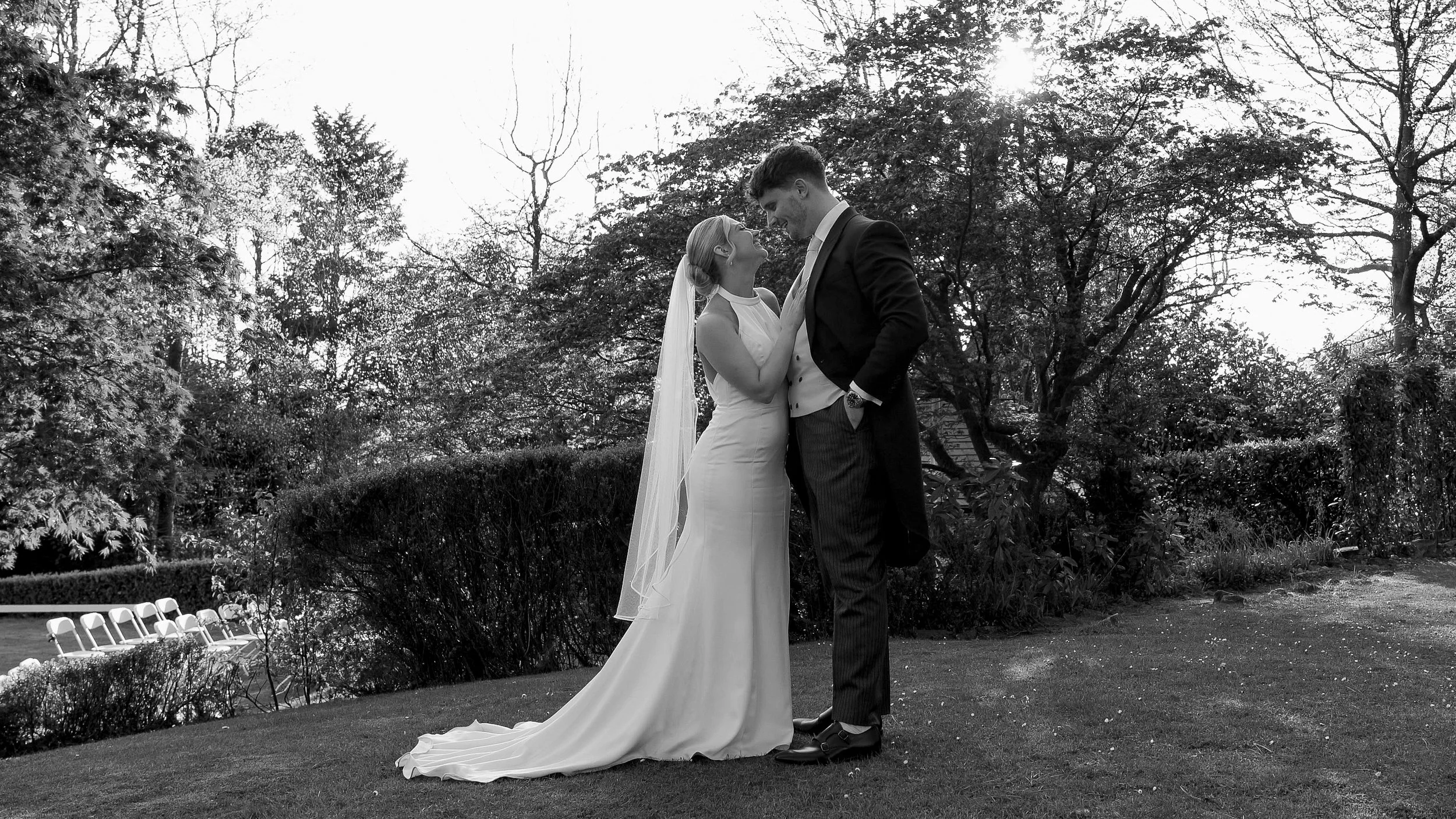 Black and white photo of a bride and groom standing outdoors in a garden, gazing into each other's eyes, with trees and bushes in the background.