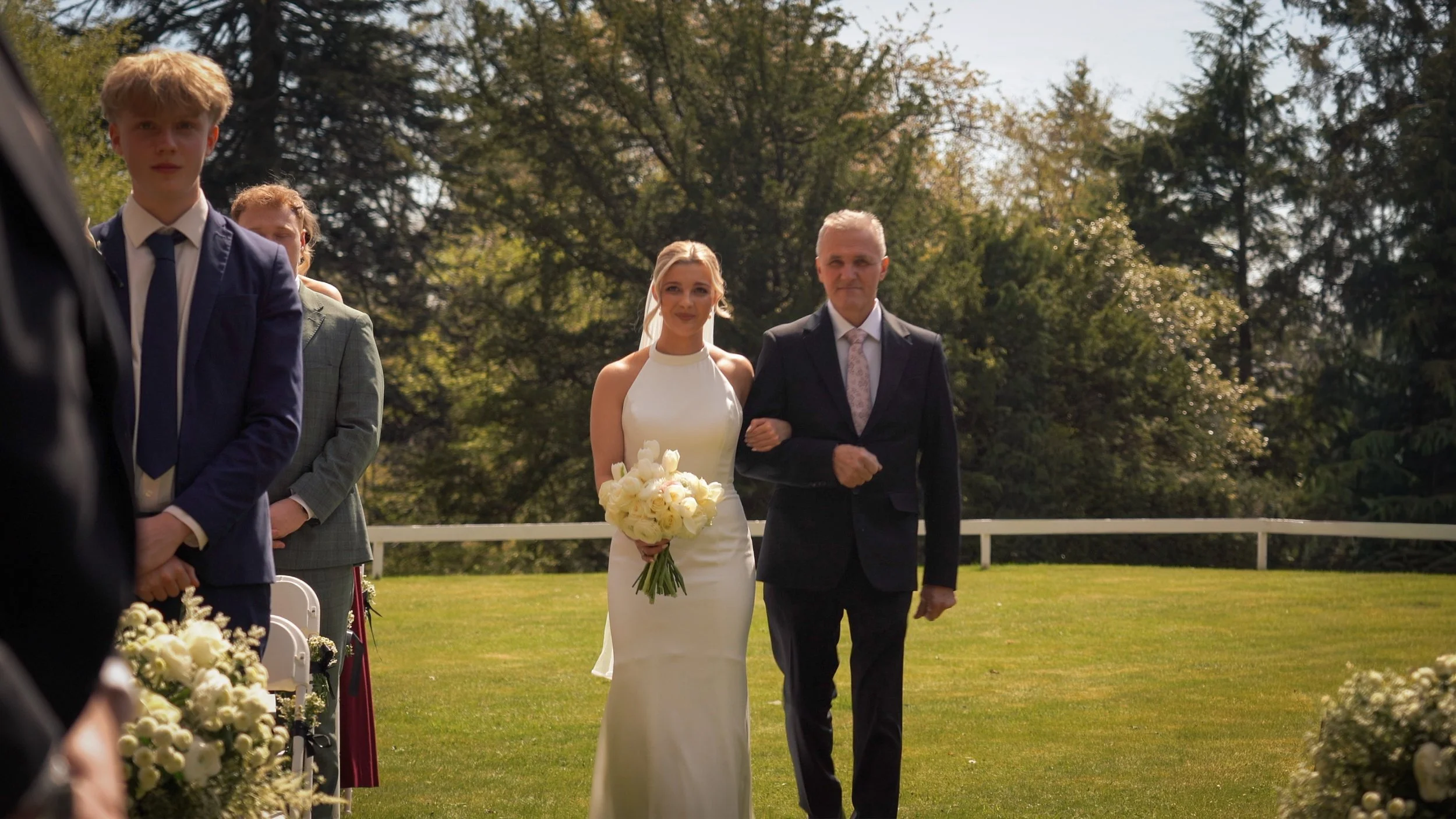 A bride in a white wedding dress holding a bouquet of white roses, walking down the aisle with an older man in a black suit, at an outdoor wedding ceremony surrounded by trees.