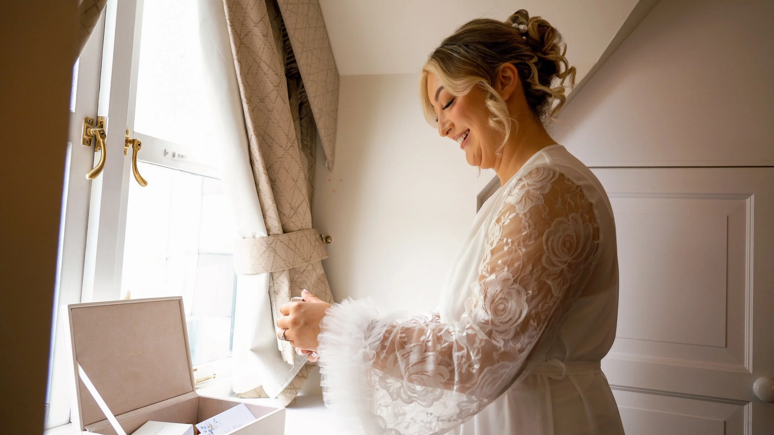 A woman with blonde hair styled in loose curls and wearing a lace robe smiles while looking down, standing near a window with beige curtains and gold handles, in a well-lit room with white walls.
