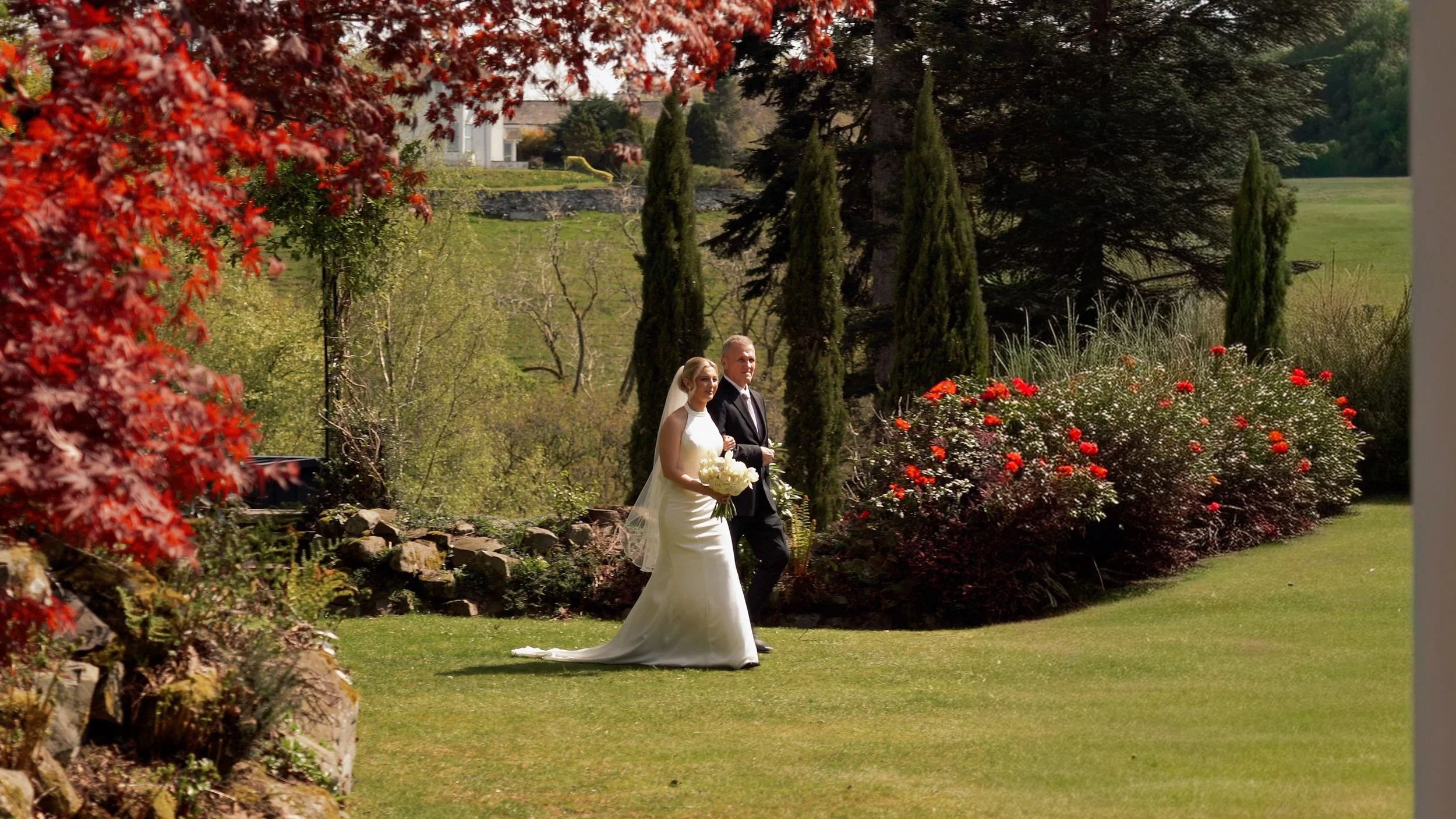 A bride and groom walking outdoors in a lush garden, surrounded by vibrant trees and flowering bushes, during a wedding.