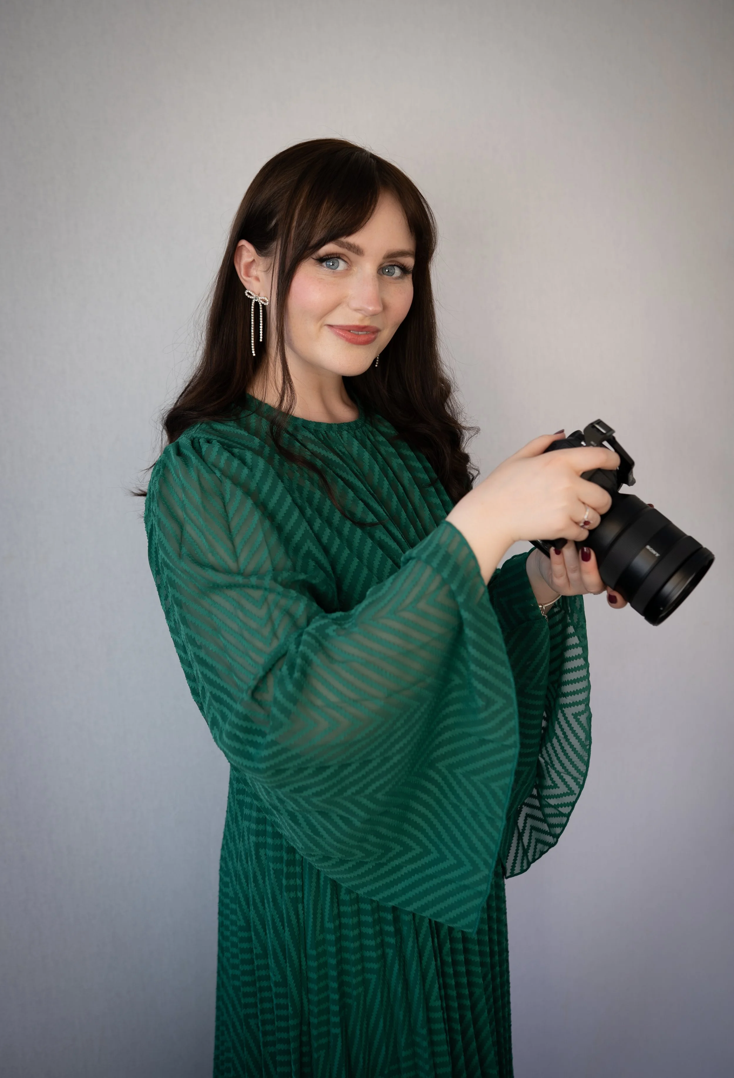 A young woman with long dark hair, blue eyes, and light makeup, wearing silver earrings and a green patterned dress, holds a camera in both hands and looks at the camera with a slight smile.