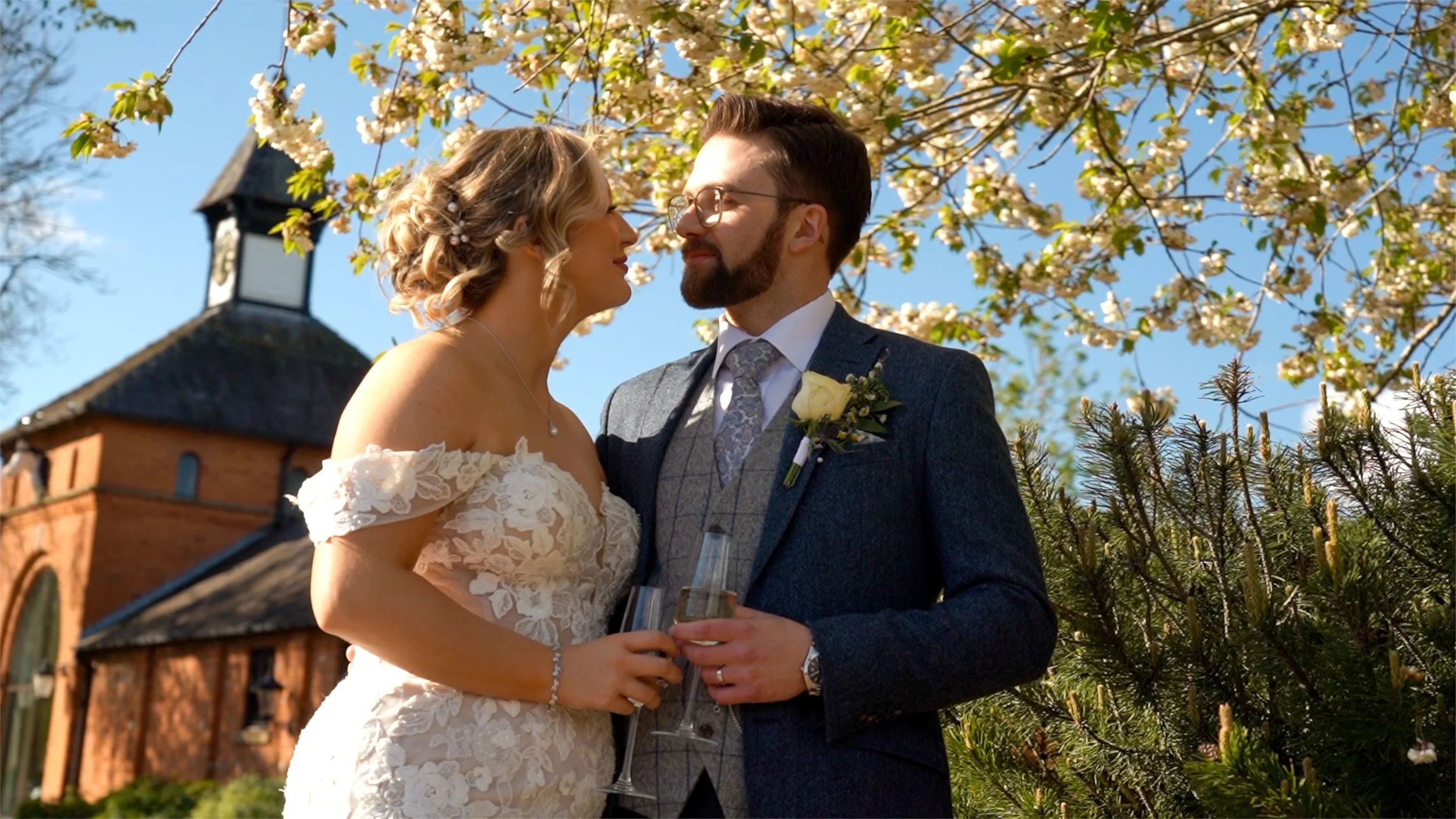 A bride and groom standing close together outdoors during their wedding, smiling and looking into each other's eyes with blooming trees and a church in the background.