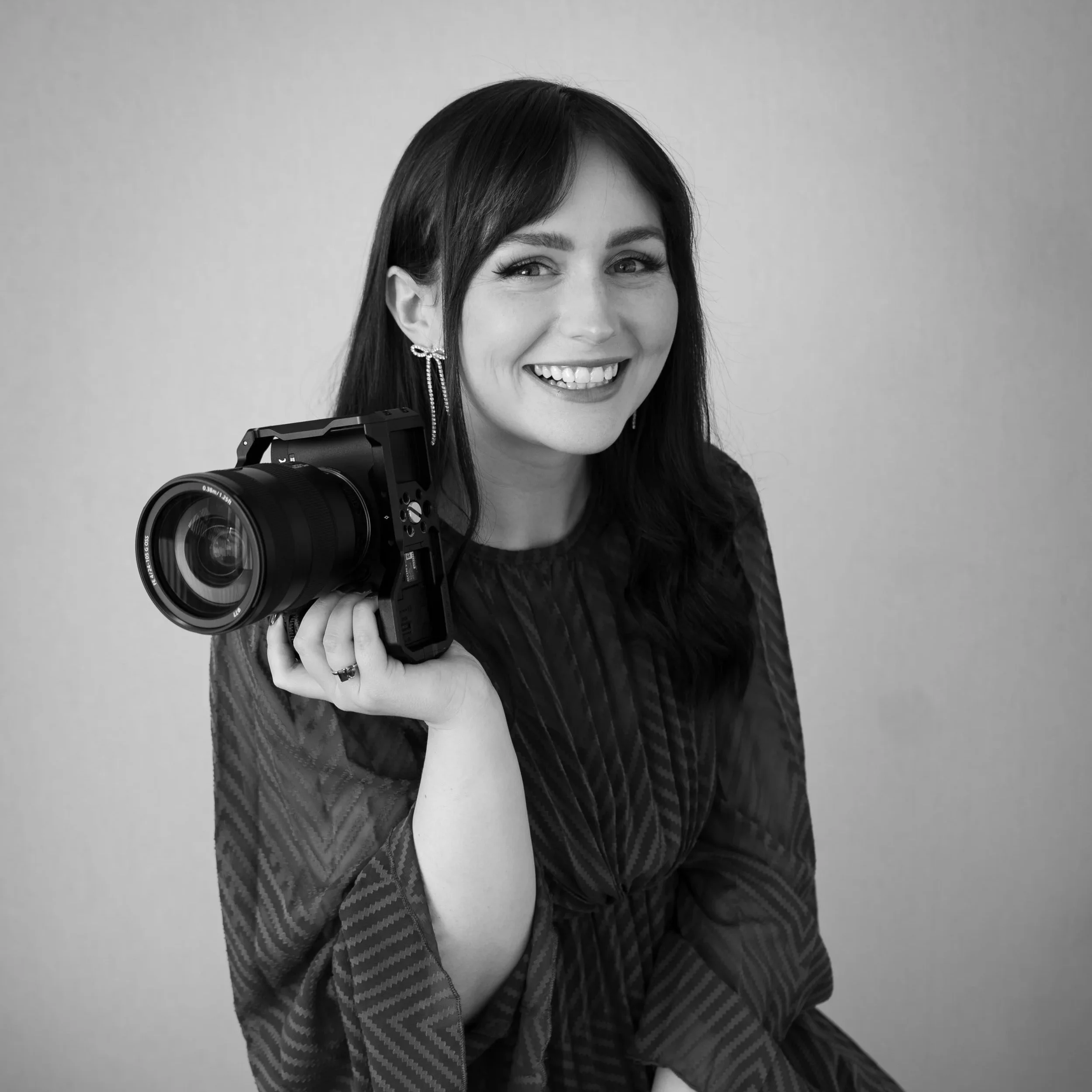 A woman with dark hair, smiling, wearing earrings and a blazer, holding a camera on her shoulder, against a plain backdrop.