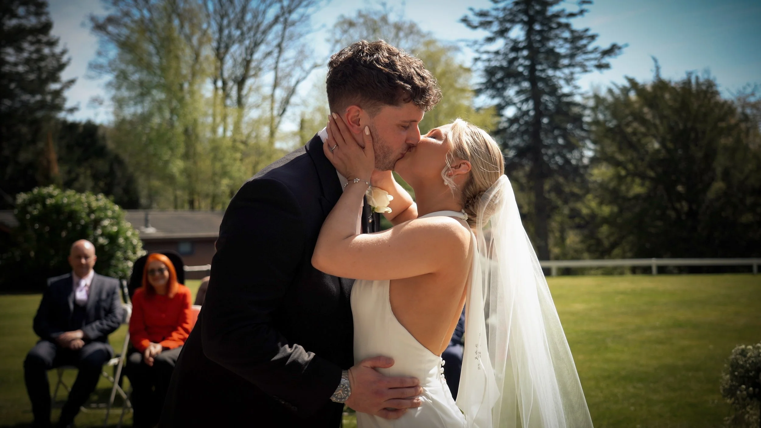 A bride and groom kissing outdoors on their wedding day, with guests seated in the background and trees surrounding the scene.