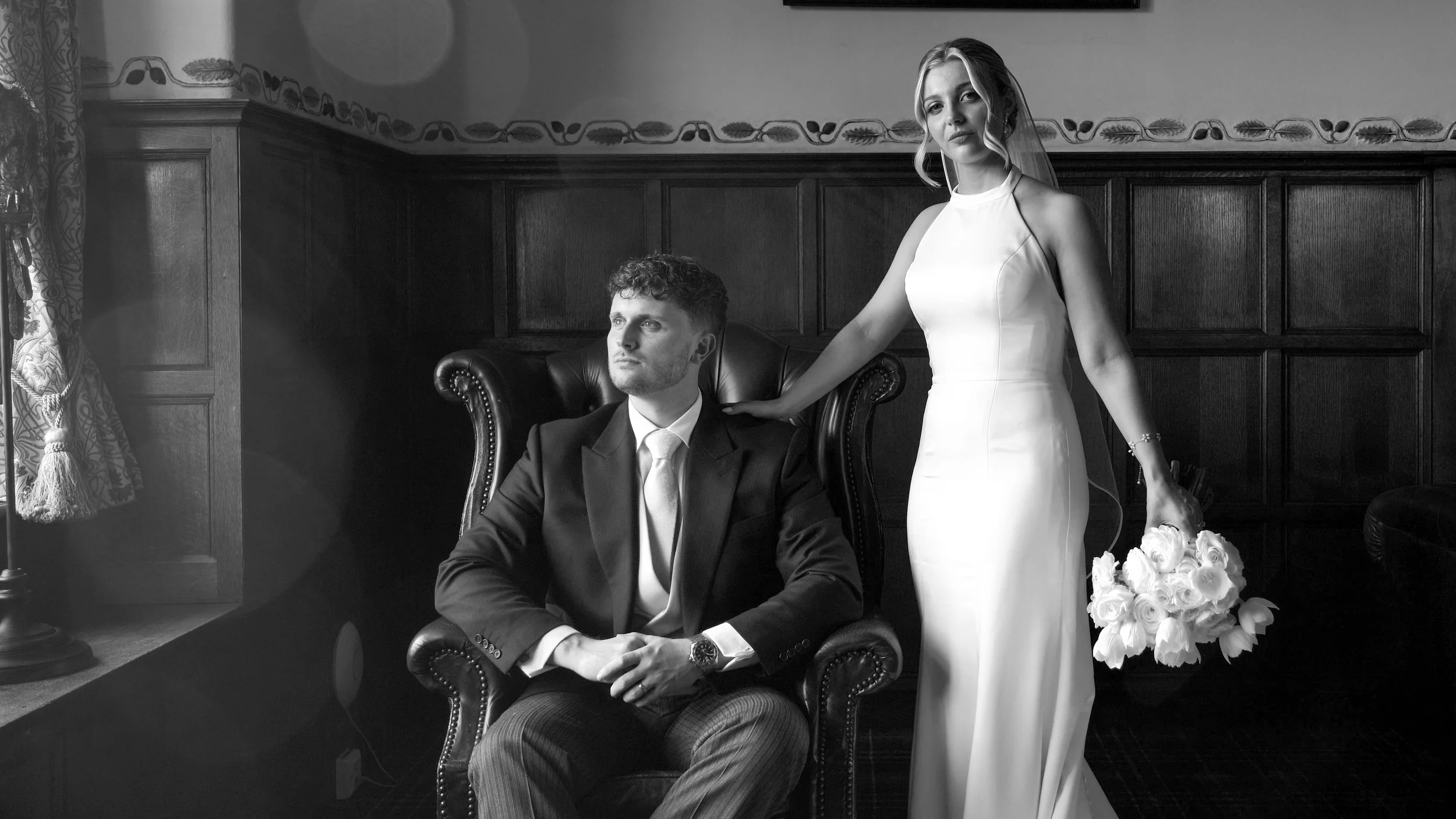 A black-and-white photo of a man in a suit seated in a leather armchair and a woman in a white gown with a bouquet of roses, standing beside him in a room with wooden paneled walls and decorative border.