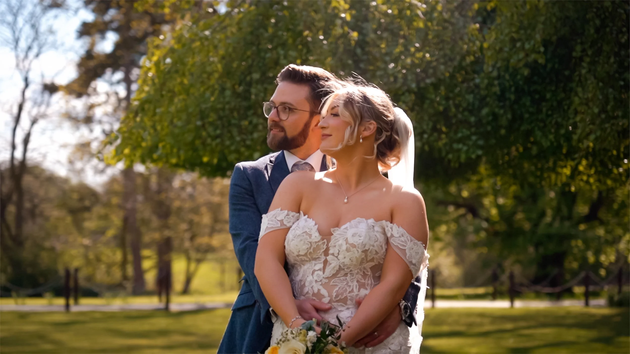 A newlywed couple outdoors, the bride in a white lace wedding dress holding a bouquet, and the groom in a suit with glasses, standing close with trees and sunlight in the background.
