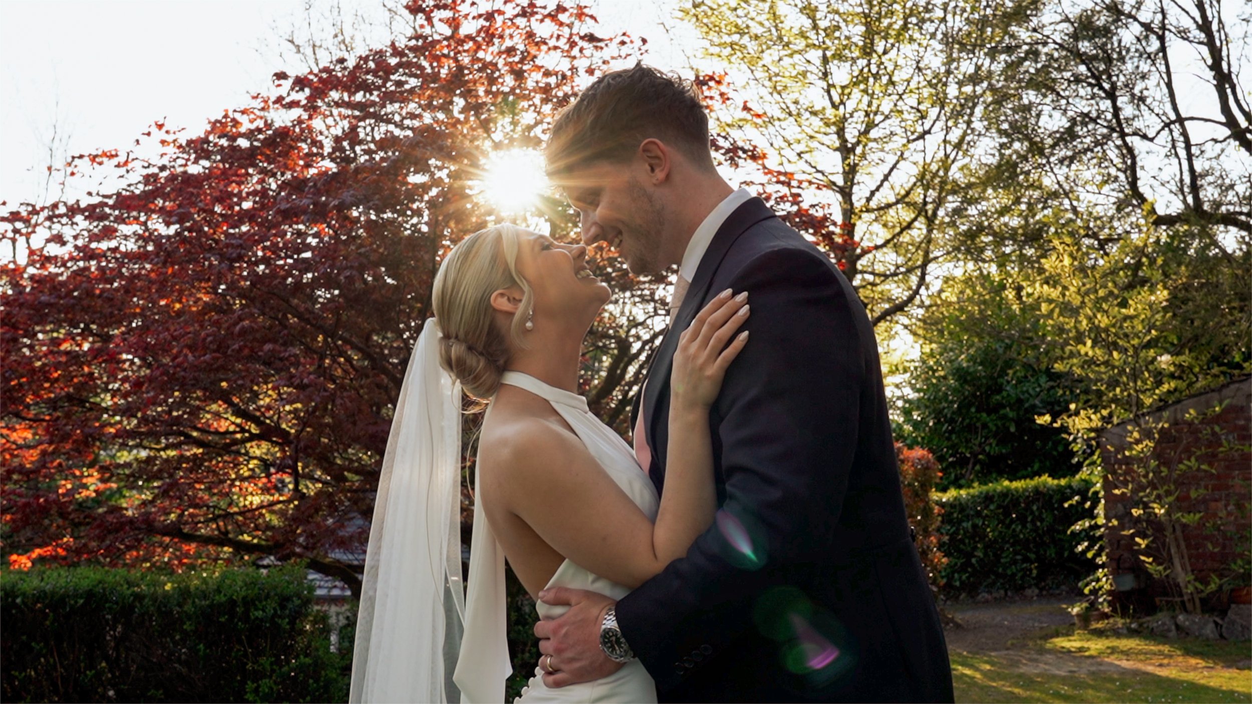 A bride and groom smiling and looking at each other while embracing during their wedding outdoors in front of trees with autumn leaves and sunlight.