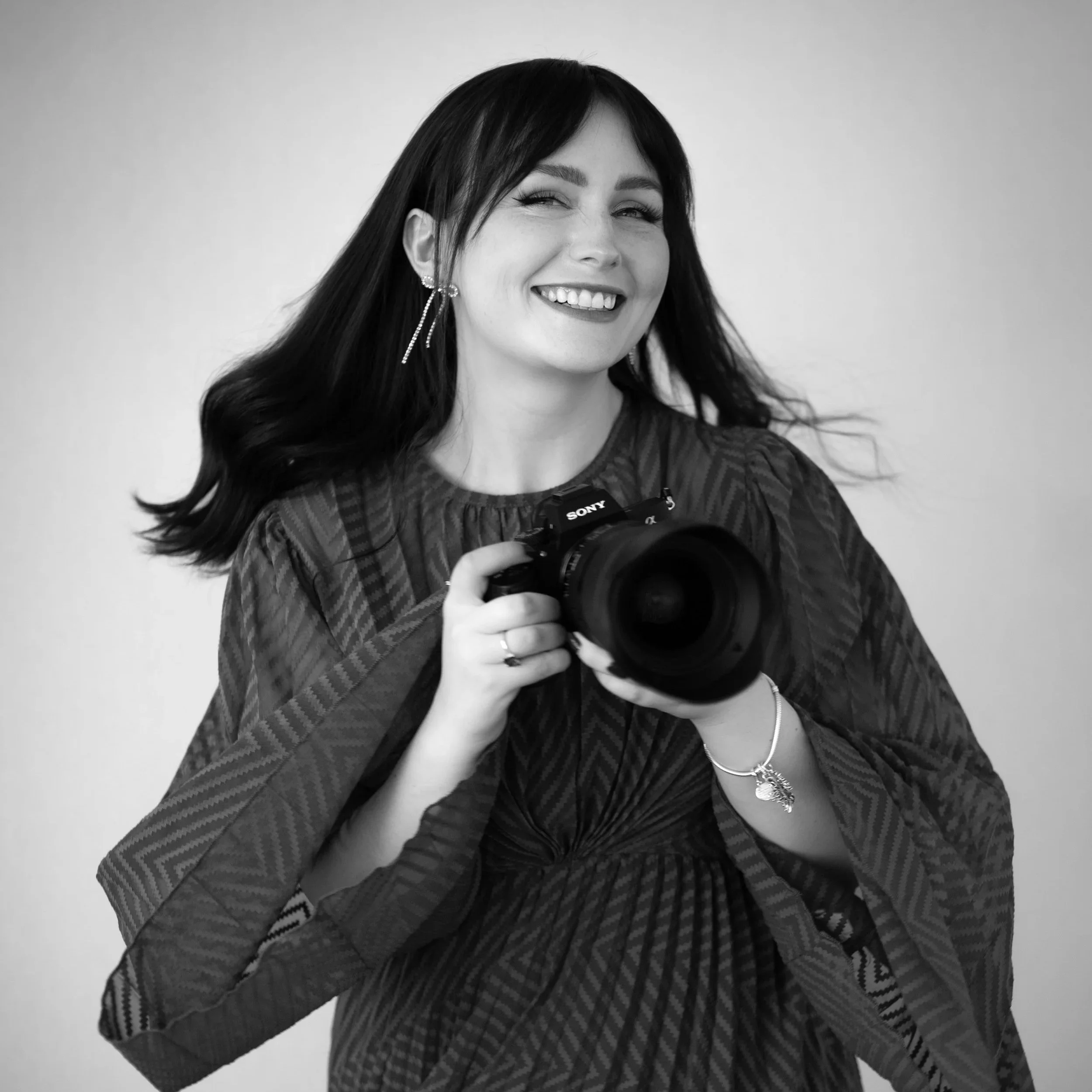 Black-and-white portrait of a woman with dark hair, smiling broadly and holding a camera, wearing earrings and a patterned sweater.