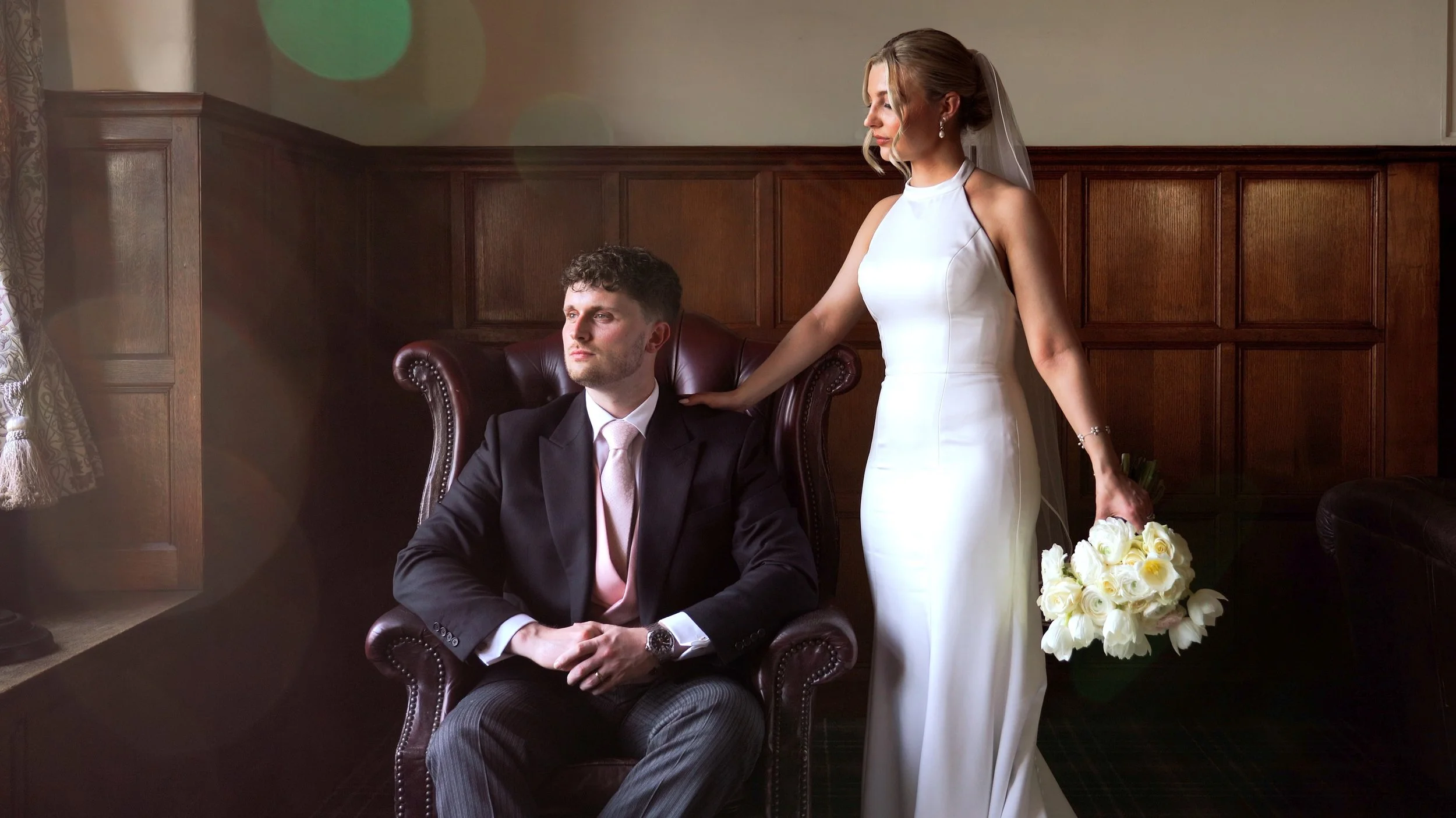 A bride in a white wedding dress standing next to a groom in a black suit and pink tie seated in a leather armchair. She is holding a bouquet of white roses and resting her hand on the groom's shoulder, with a wooden-paneled wall behind them.