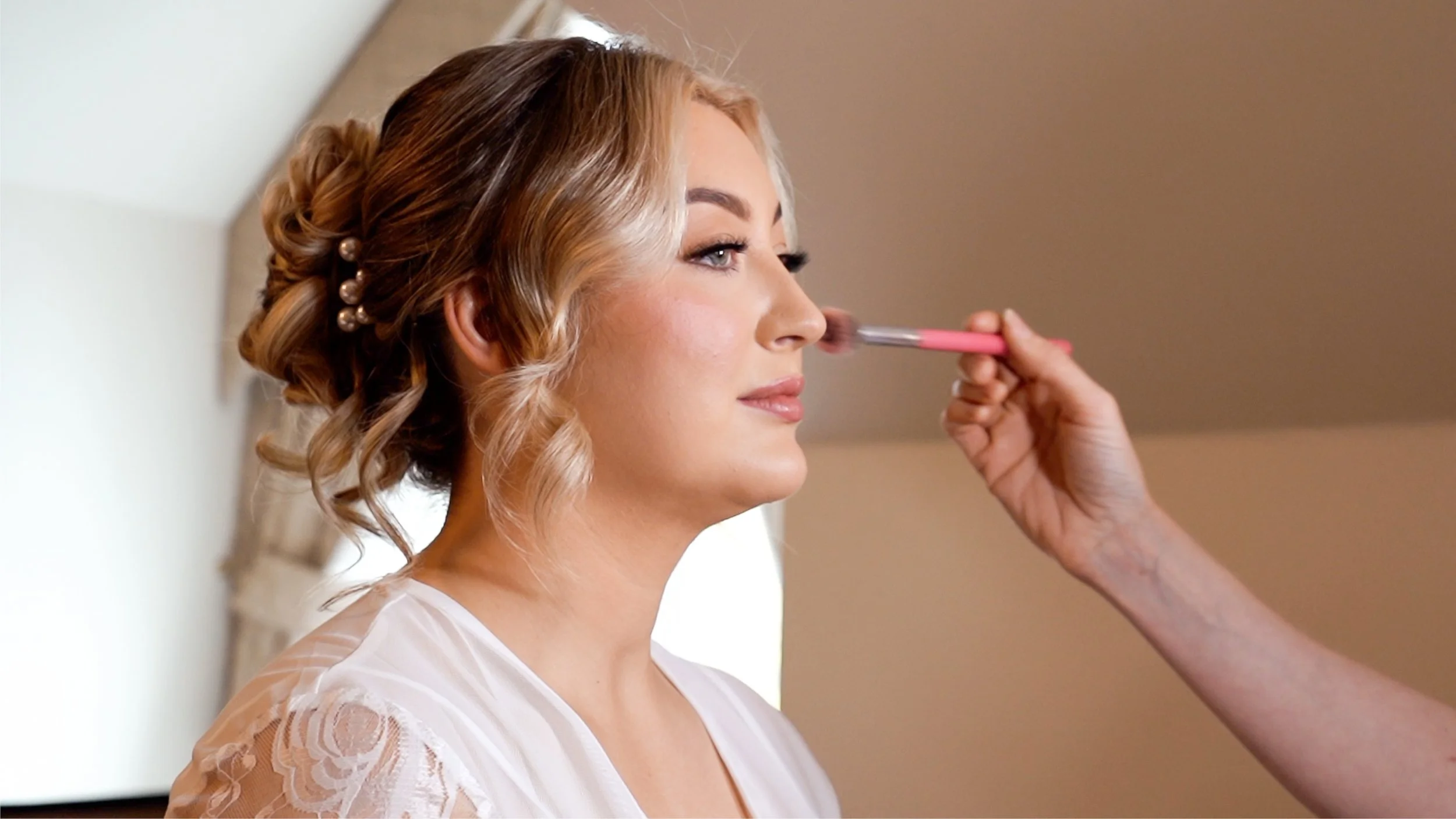 A woman with styled blonde hair is having makeup applied to her face by a makeup artist. The woman is wearing a white dress with lace details on the shoulder.