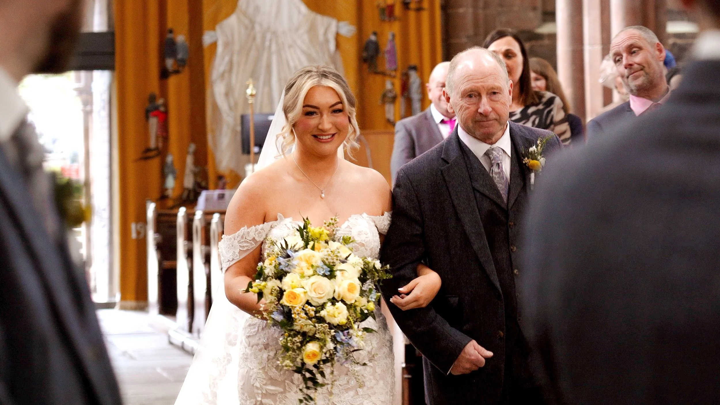 A bride in a wedding dress holding a bouquet of yellow, white, and purple flowers, walking down the aisle with an older man in a suit during a wedding ceremony inside a church.