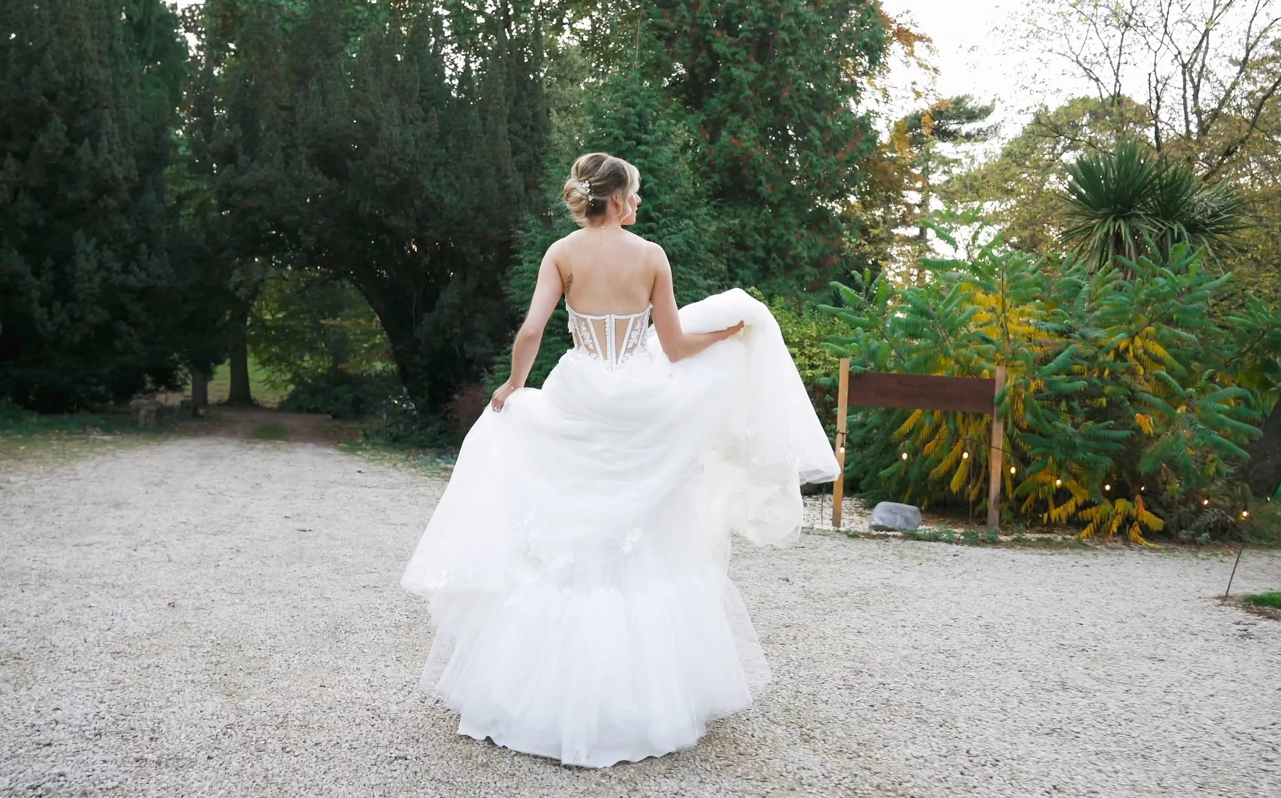 A bride in a white wedding dress sitting on a ledge outdoors surrounded by green trees.