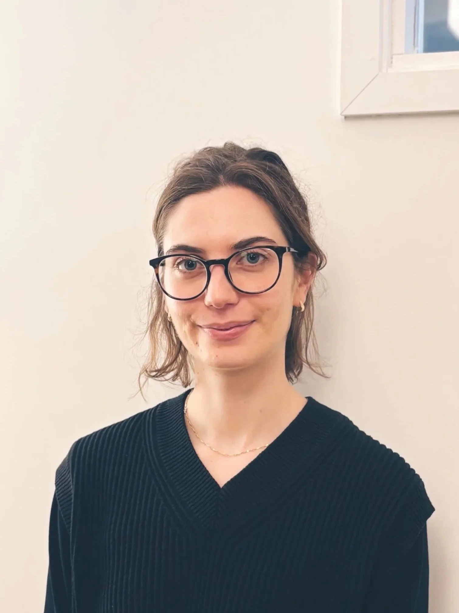 A woman with glasses and short brown hair, smiling and wearing a black top, standing against a plain white wall with a window in the background.