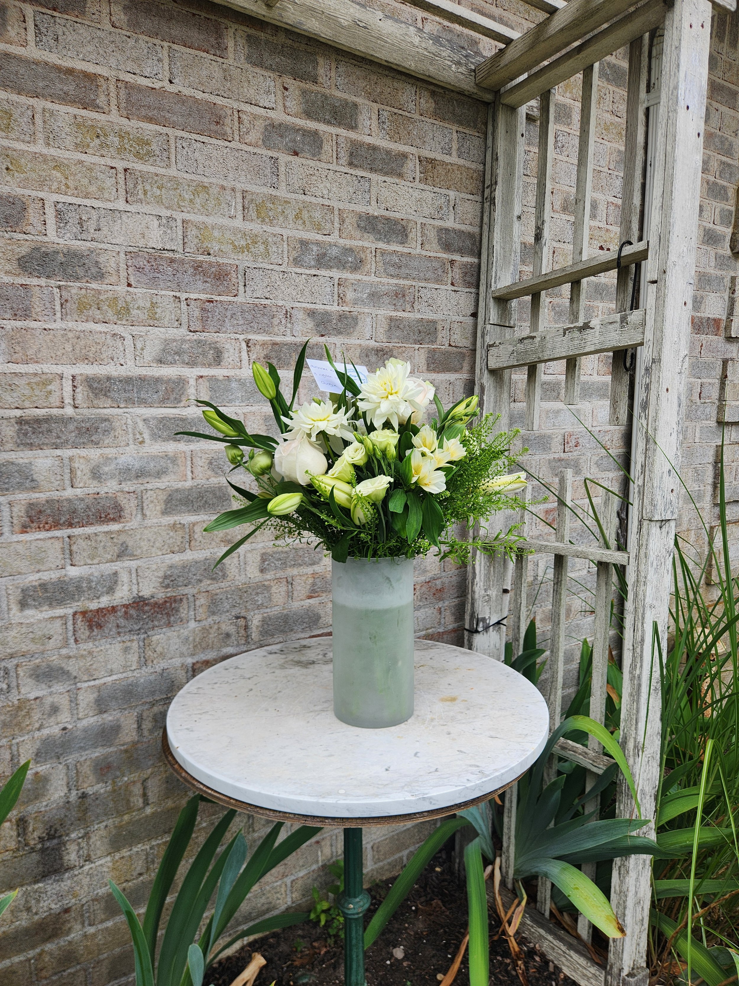 A white marble round table with a green metal base in a garden. On top of the table is a gray vase holding white lilies and greenery. Behind the table, there is a brick wall and a weathered wooden trellis with some green plants around.