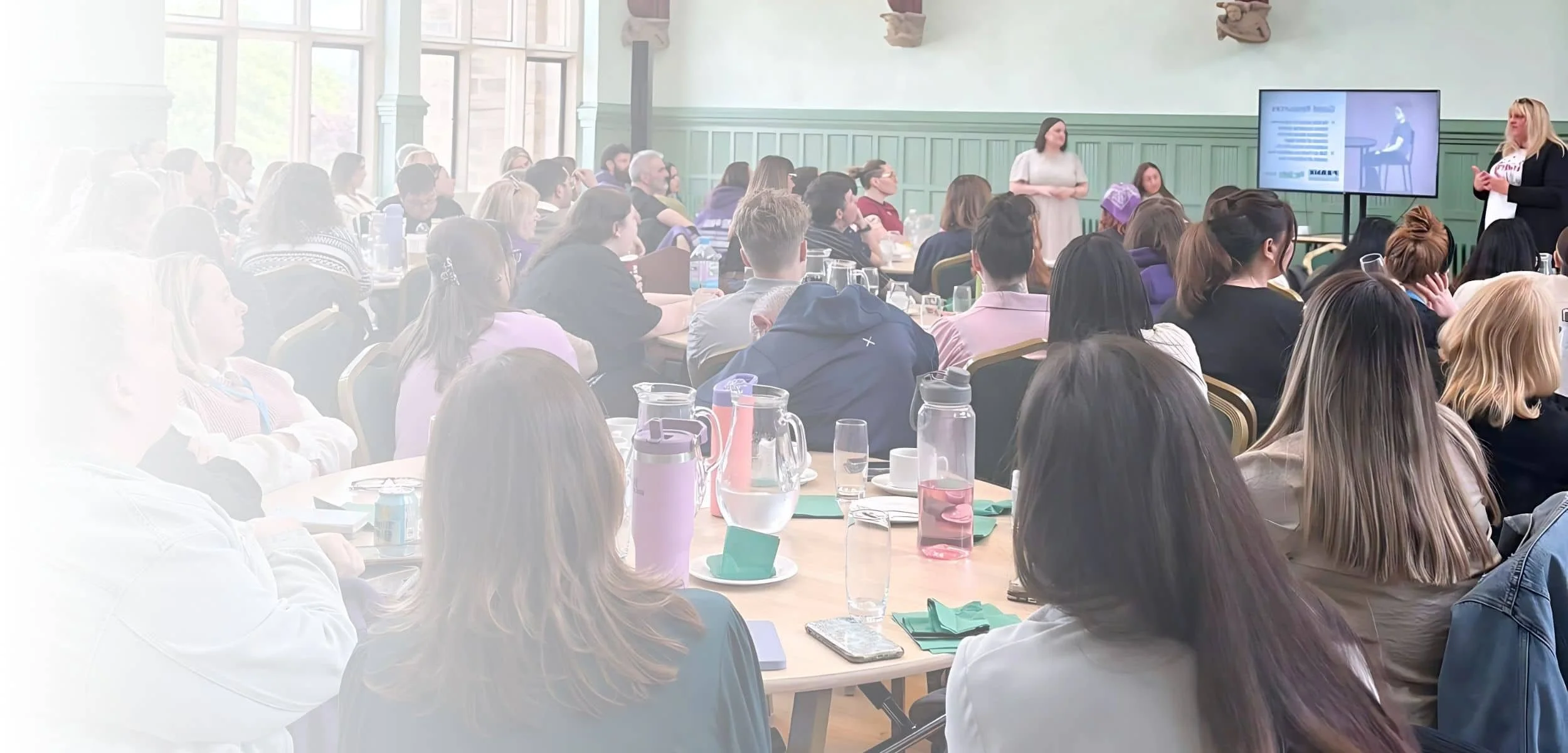 A large group of people attending a conference or seminar in a bright, spacious room with green wall paneling and large windows. A woman is presenting at the front, next to a screen displaying a presentation slide.
